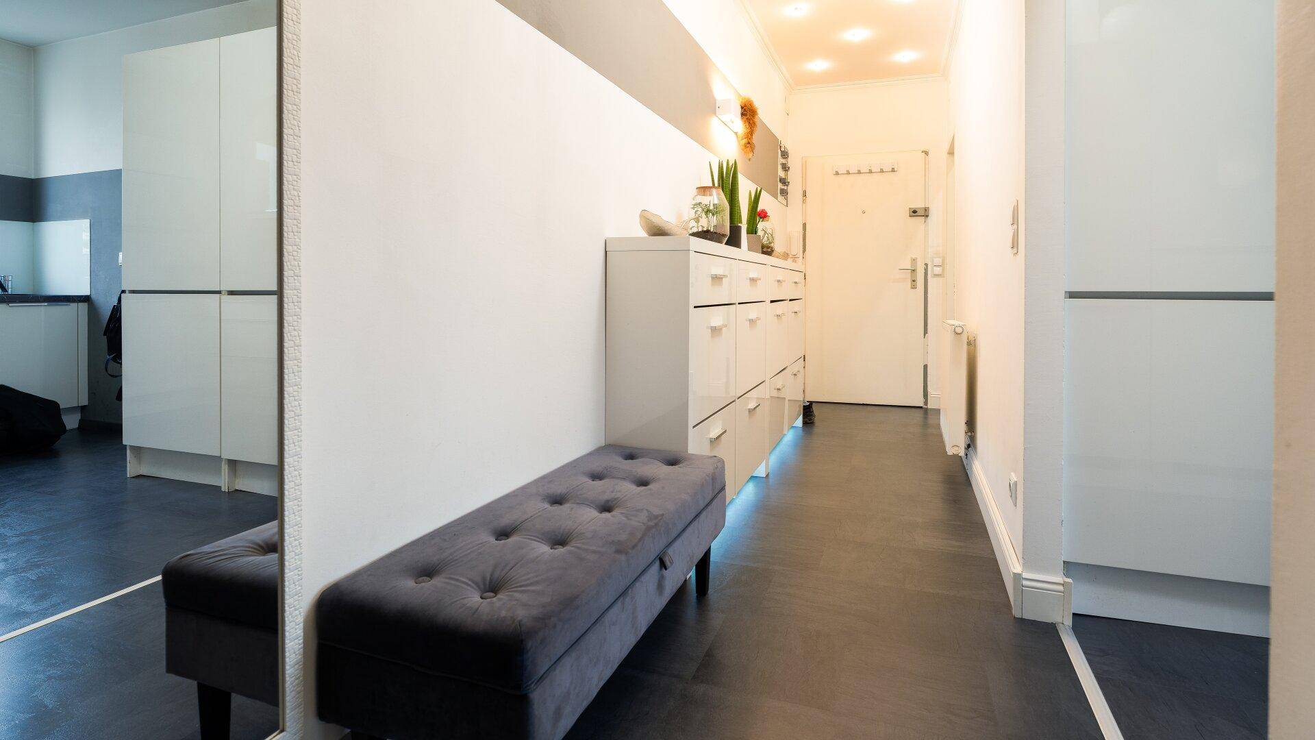 Langer Flur mit dunklem Boden, einem Spiegel und einer Sitzbank, die zum Eingangsbereich führt.