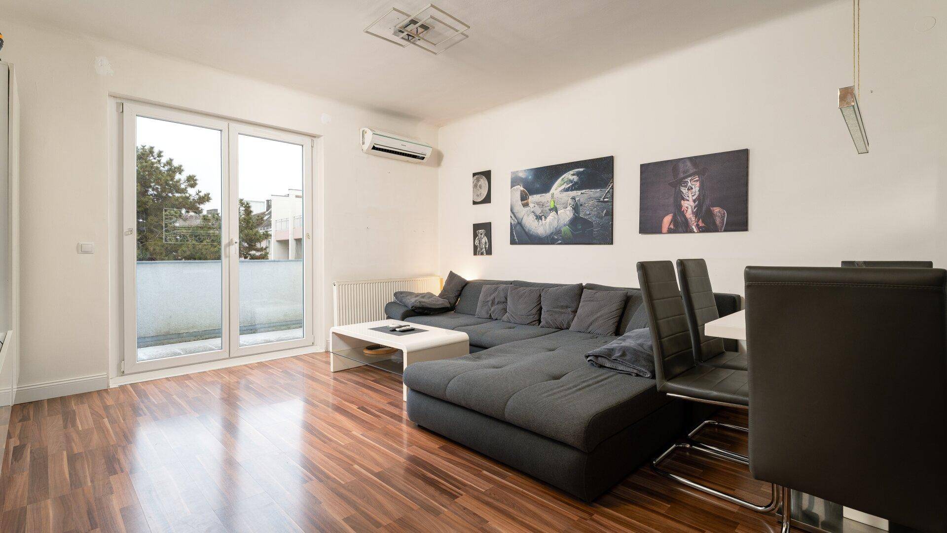 Heller Wohn- und Essbereich mit Holzboden, großem Sofa und Zugang zu einem Balkon.