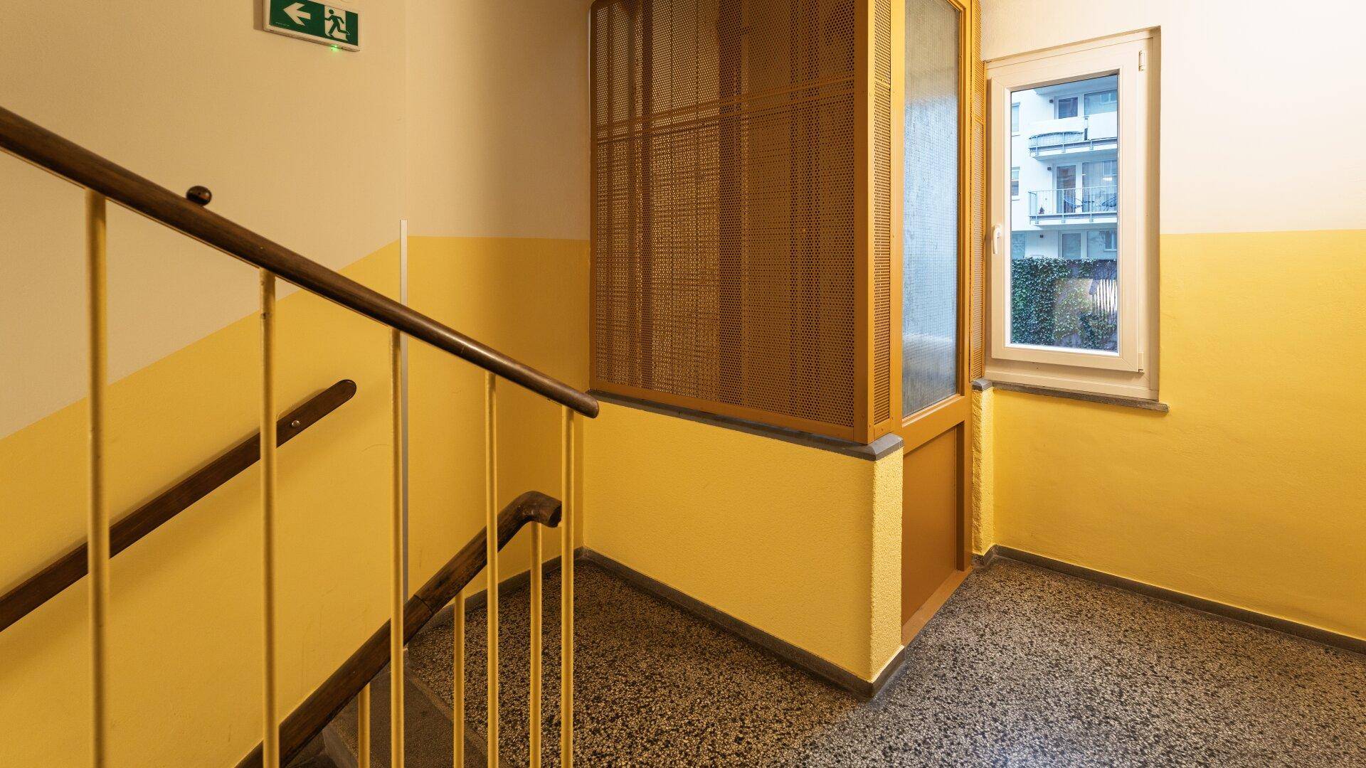 Treppenhaus mit gelb gestrichenen Wänden, Terrazzoboden und einem Aufzug im Hintergrund.