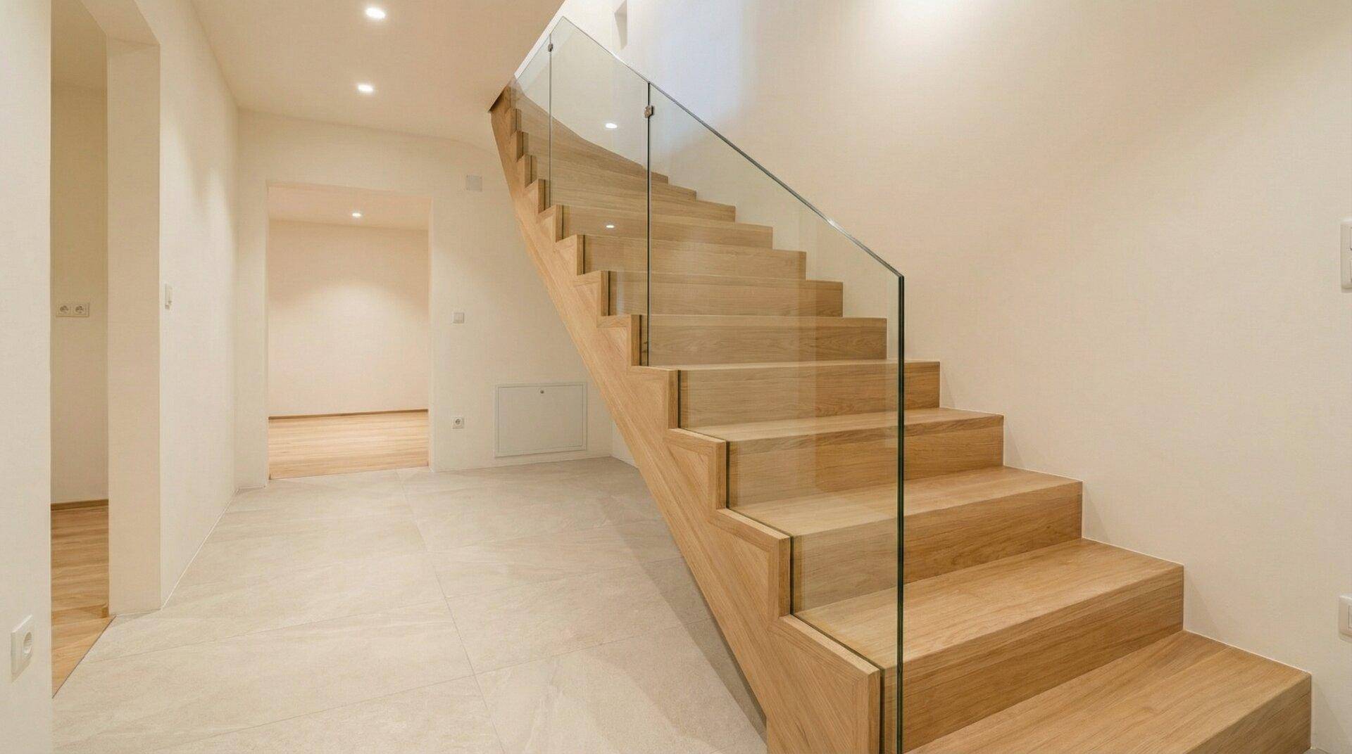 Heller Flur mit moderner Holztreppe, Glasgeländer und Fliesenboden im Erdgeschoss.