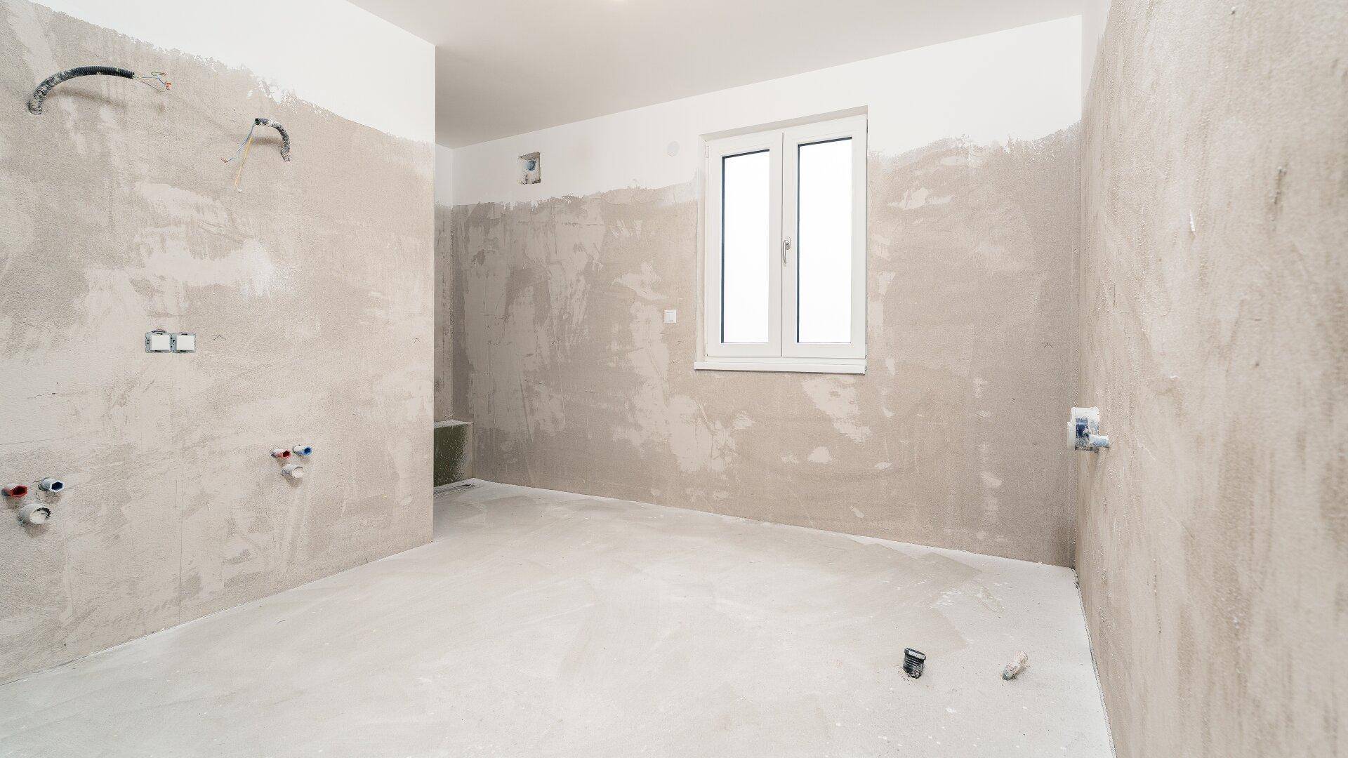 Raum im Rohbauzustand mit sichtbaren Installationen für Küche oder Bad und Fenster.