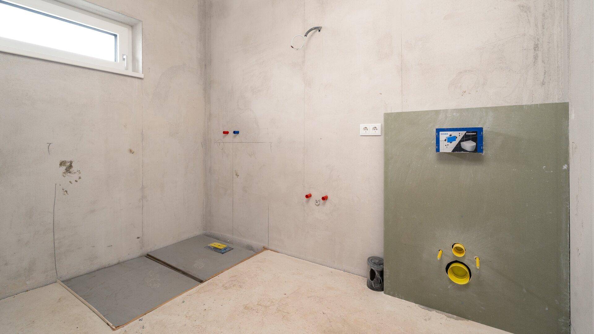 Unfertiges Badezimmer im Rohbau mit sichtbaren Installationen und Fenster.