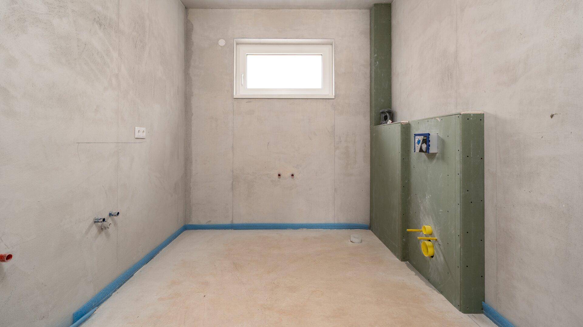 Unfertiges Badezimmer im Rohbau mit sichtbaren Installationen und Fenster.