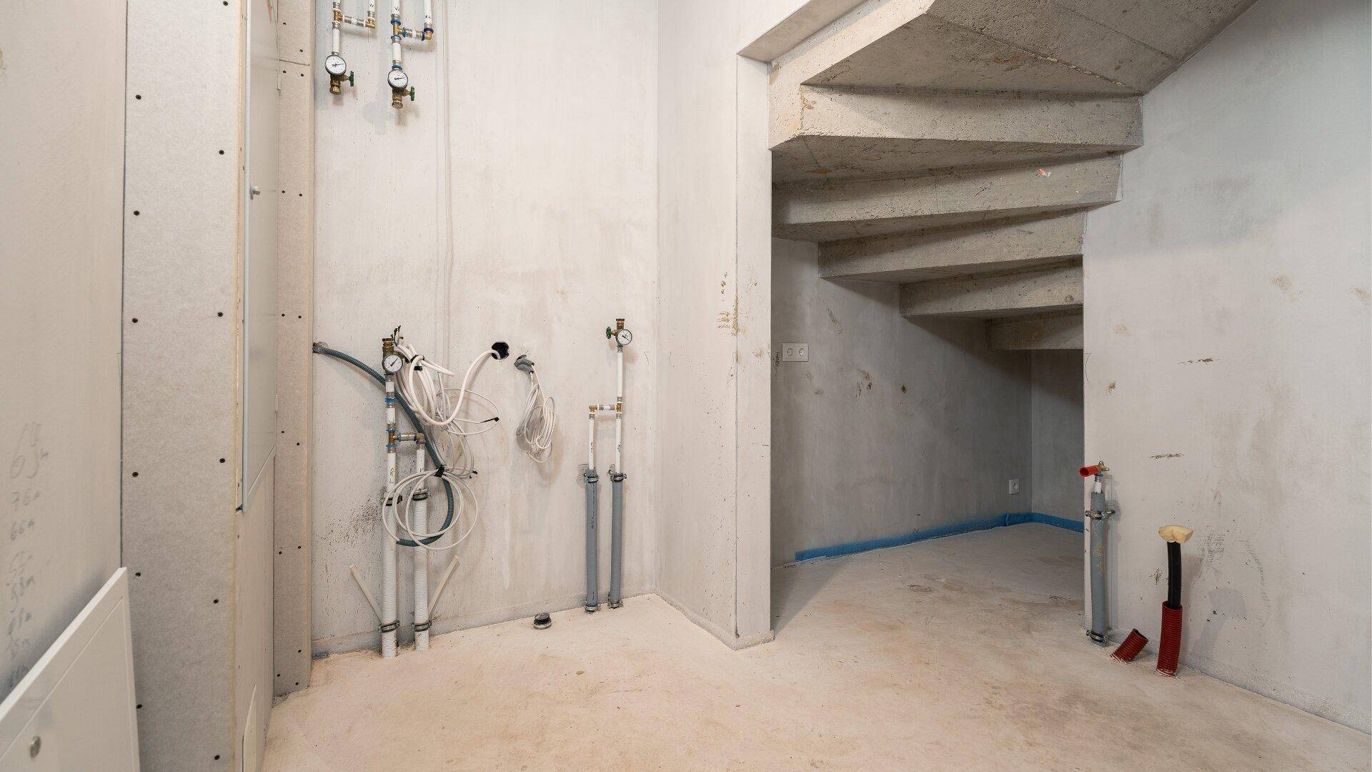 Unfertiger Kellerraum mit sichtbaren Installationen und Betontreppe im Rohbau.