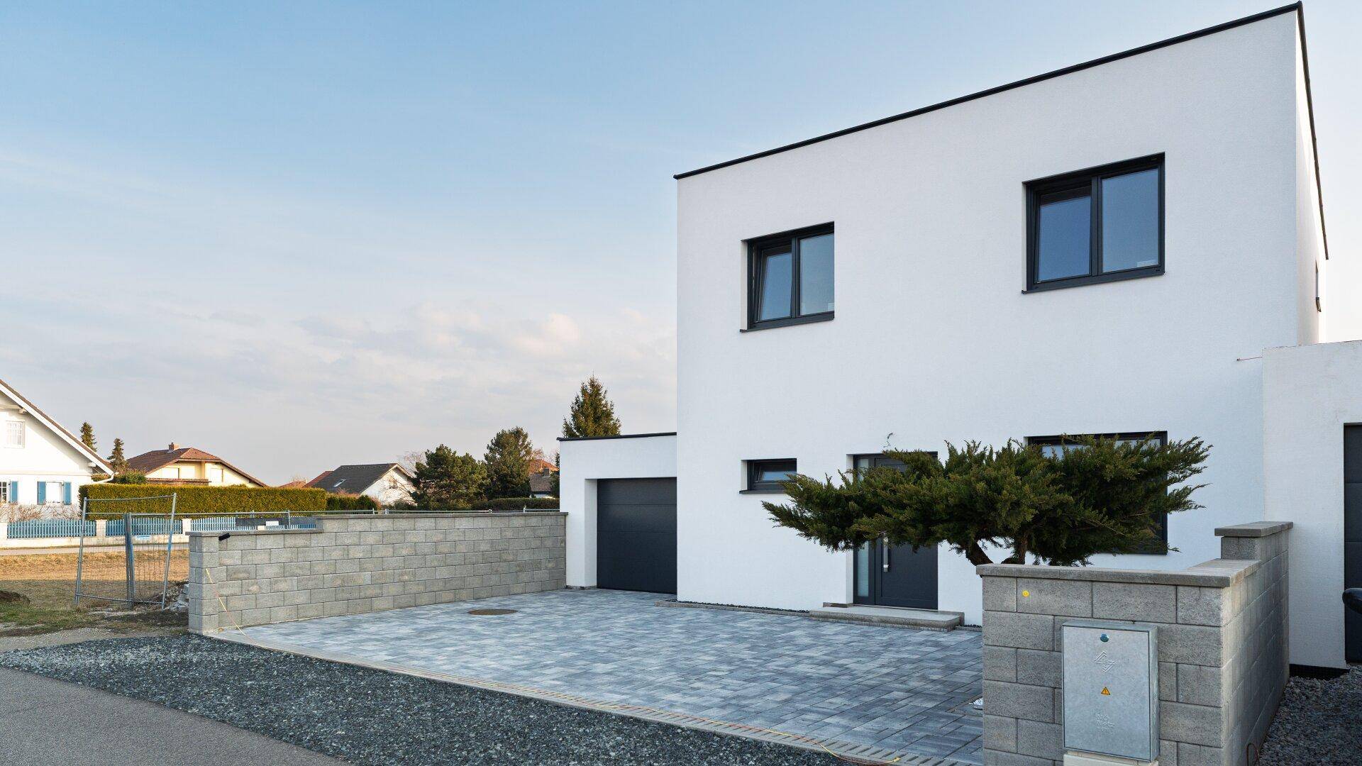 Ansicht des modernen Einfamilienhauses mit weißer Fassade, Garage und gepflastertem Vorplatz.