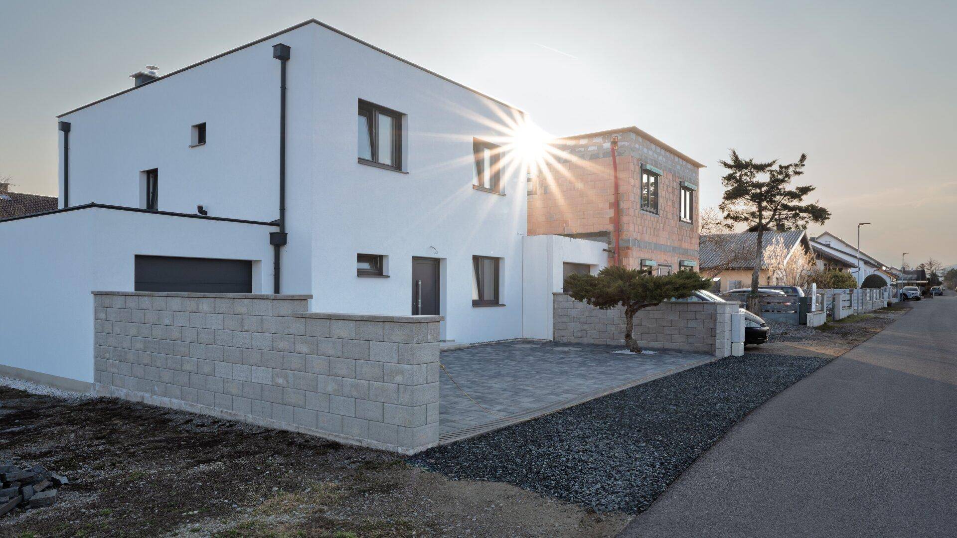 Seitenansicht des modernen Neubaus mit weißer Fassade und angrenzendem Rohbau bei Sonnenuntergang.