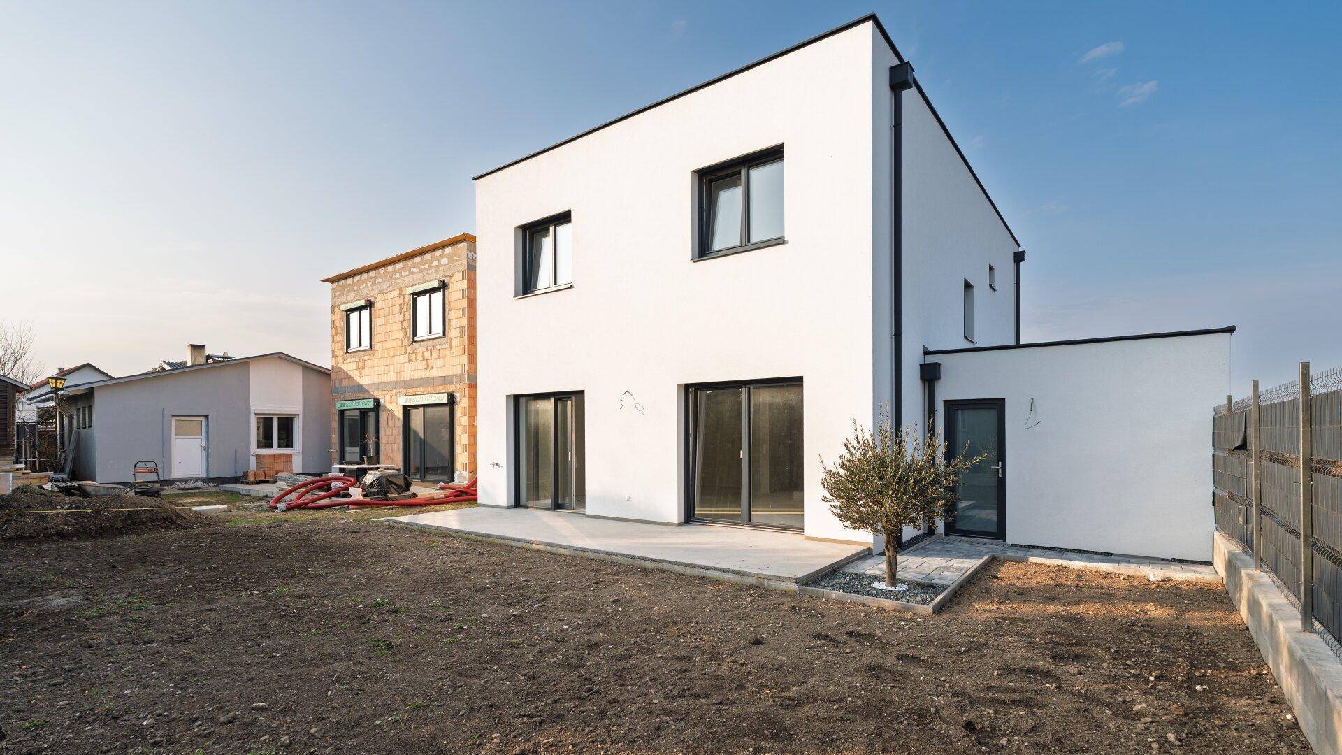 Seitenansicht des modernen Neubaus mit weißer Fassade, angrenzendem Rohbau und unfertigem Gartenbereich.