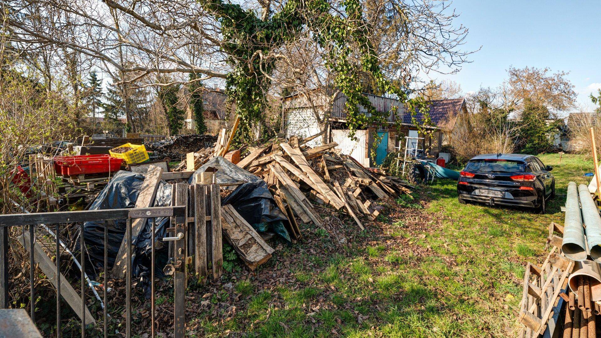 Verwilderter Gartenbereich mit Holzstapeln, Schuppen und einem geparkten Auto.