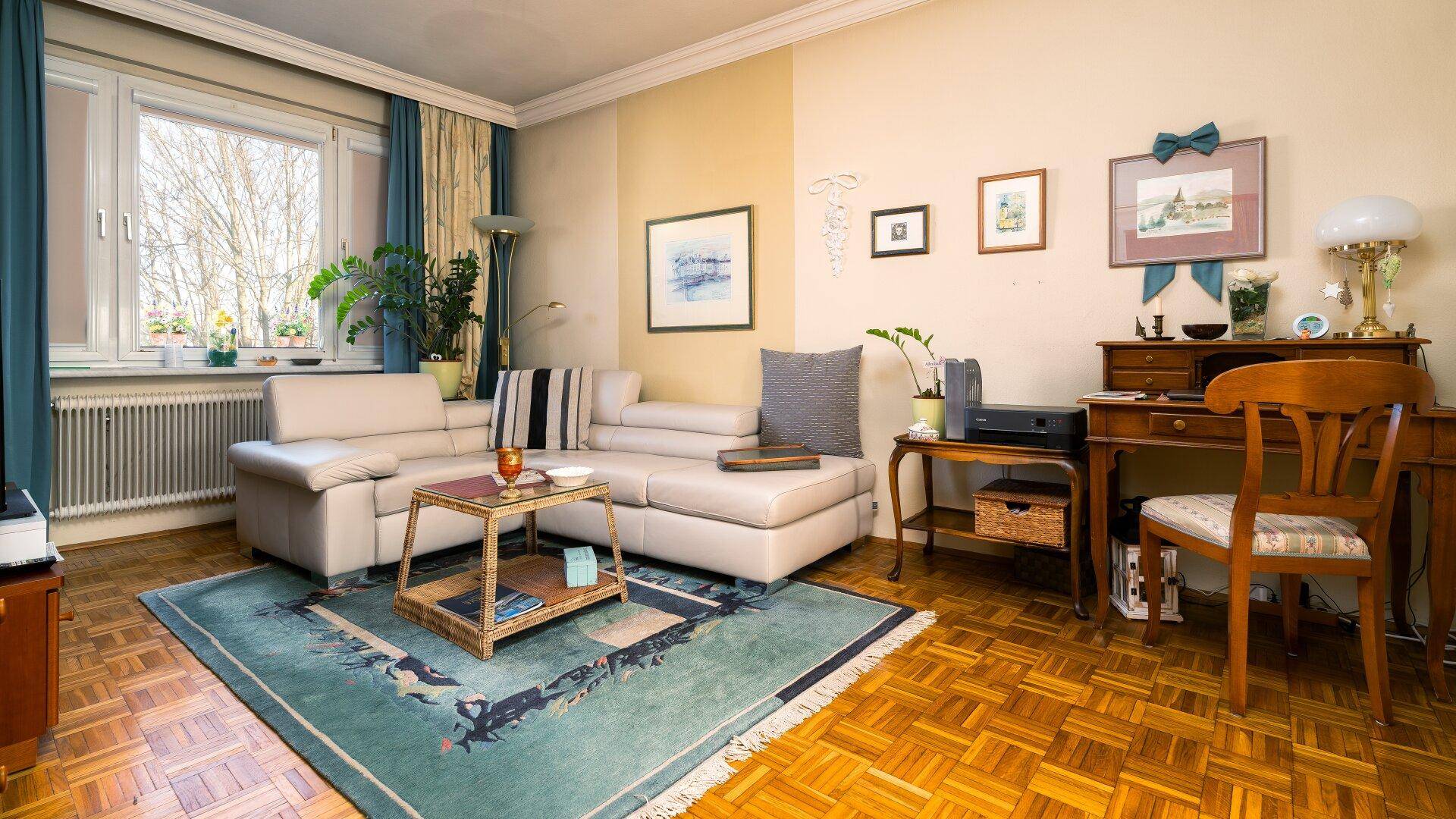 Helles Wohnzimmer mit beiger Eckcouch, Parkettboden und einem kleinen Schreibtischbereich.