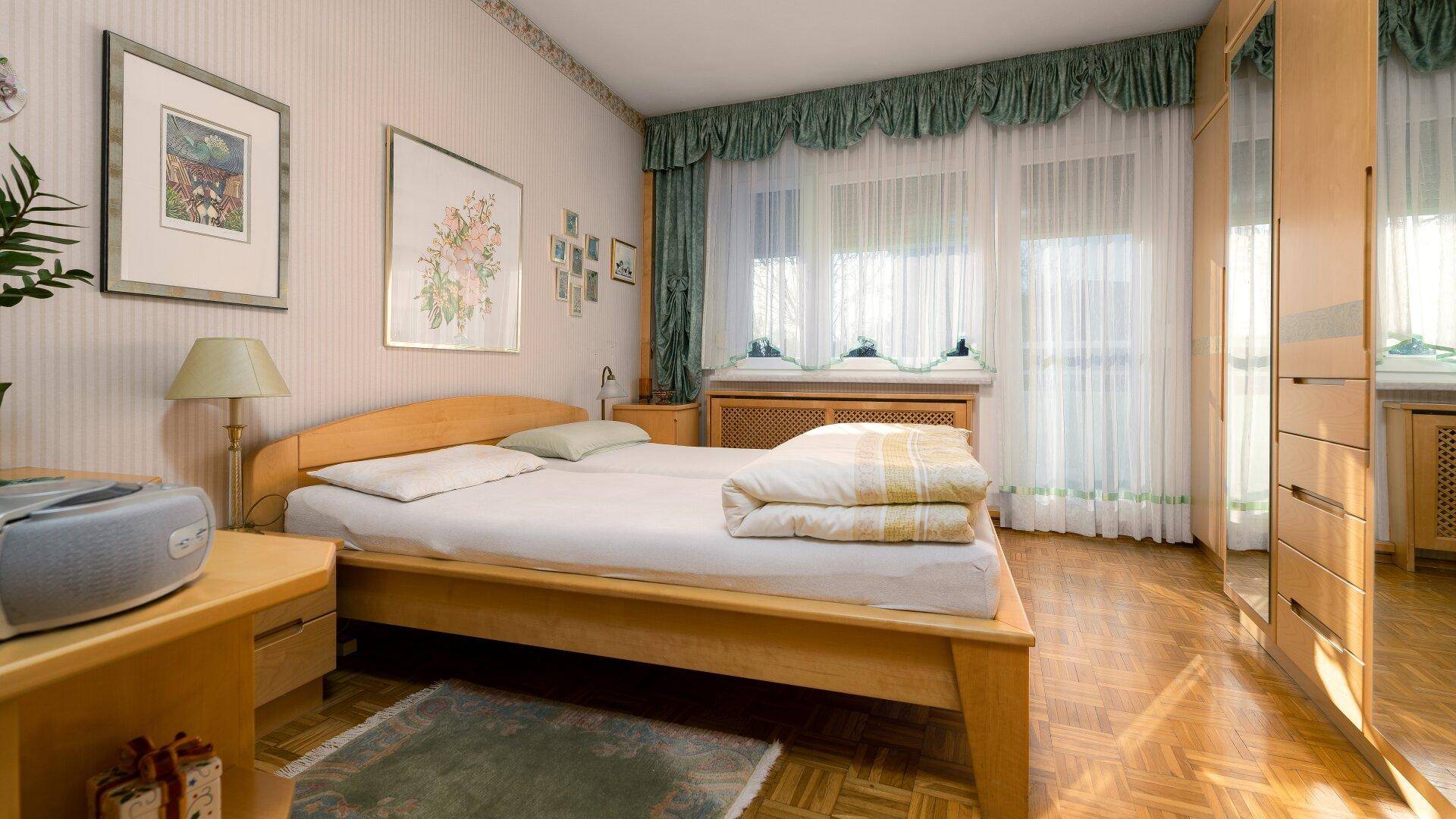 Schlafzimmer mit hellem Holzdoppelbett, Parkettboden und großen Fenstern mit Vorhängen.