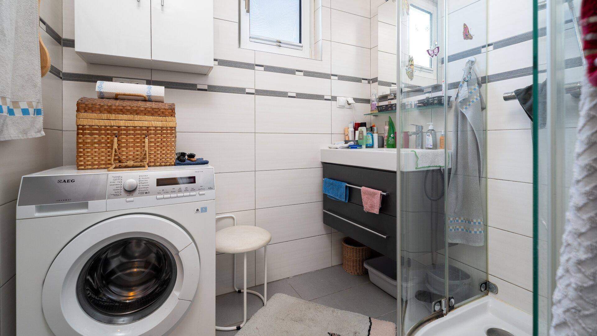 Modernisiertes Badezimmer mit Dusche, Waschbecken, Waschmaschine und weißen Fliesen.