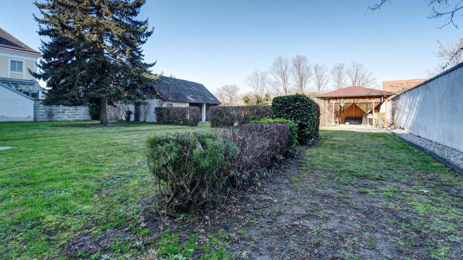 Großer Gartenbereich mit Rasenfläche, Hecken und einem Gartenhaus im Hintergrund.
