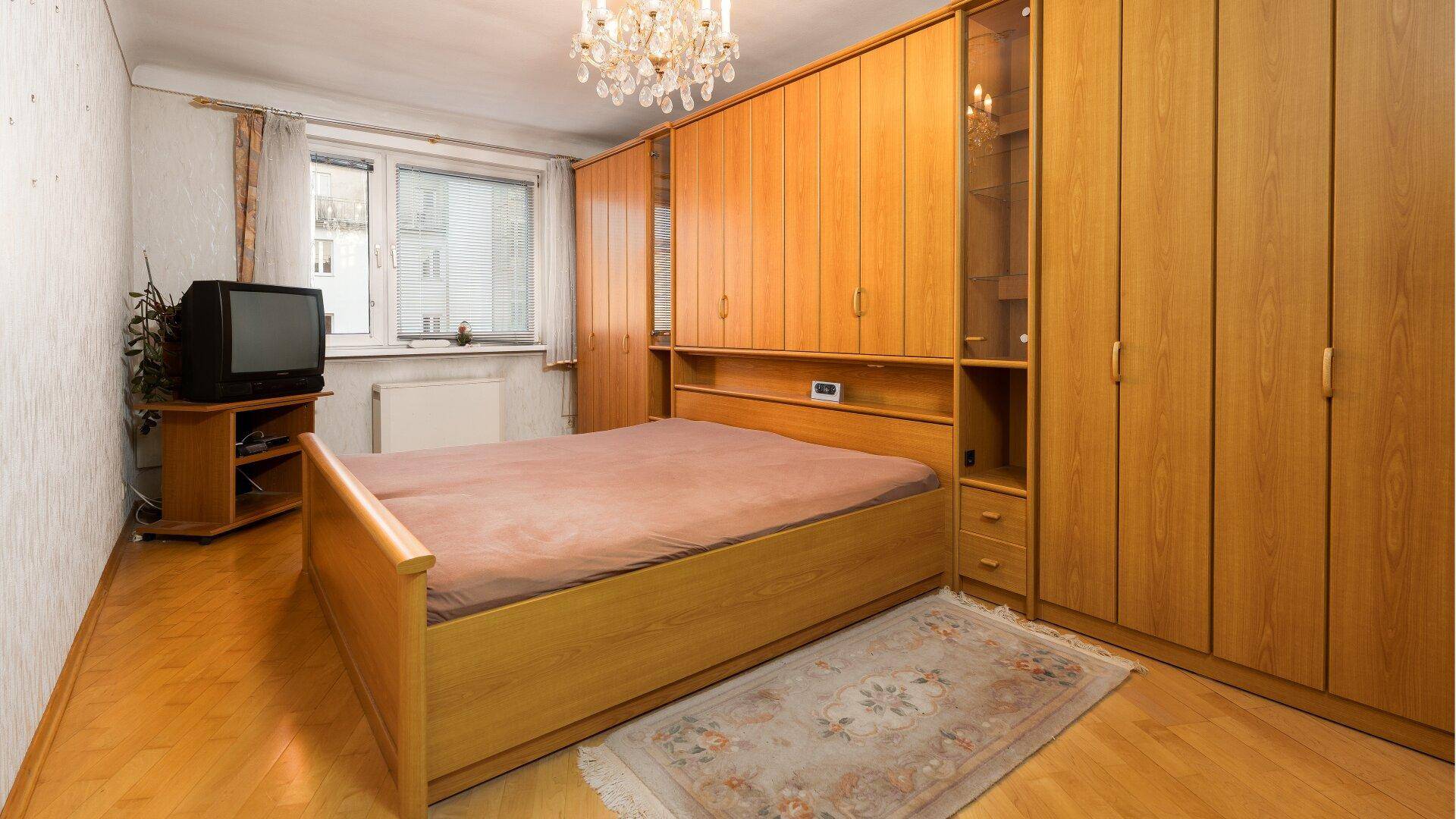 Schlafzimmer mit hellem Holzbett, großem Einbauschrank und Parkettboden, gut beleuchtet durch ein Fenster.