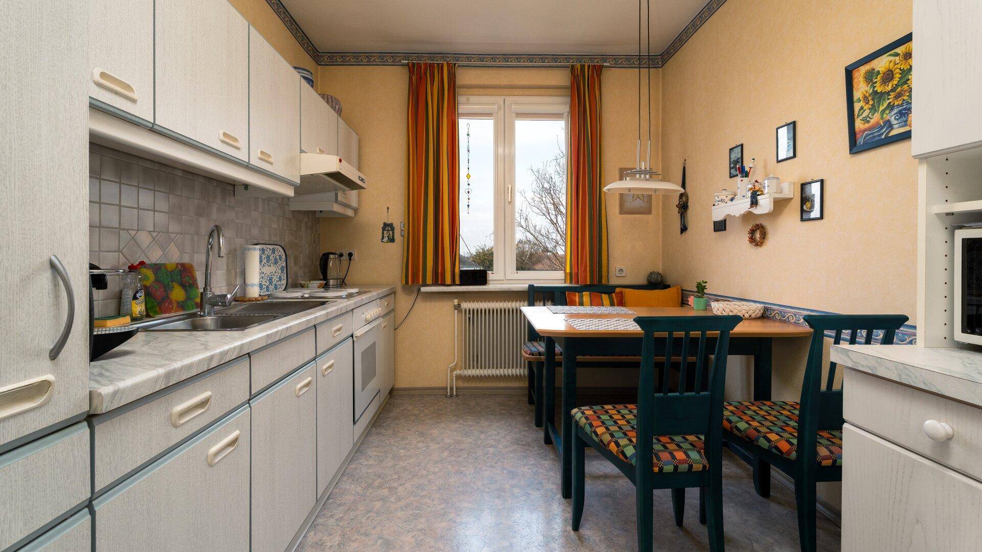 Gemütliche Wohnküche mit Esstisch, hellen Schränken und einem Fenster mit bunten Vorhängen.