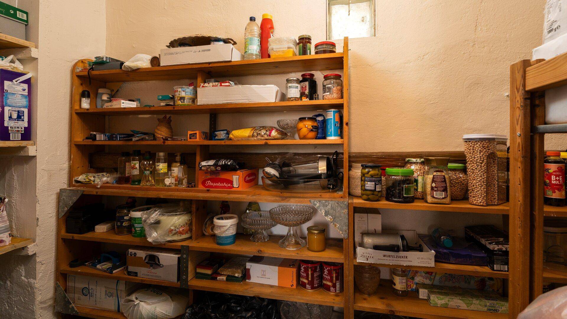 Gut gefüllte Speisekammer mit Holzregalen und zahlreichen Vorräten und Haushaltsartikeln.