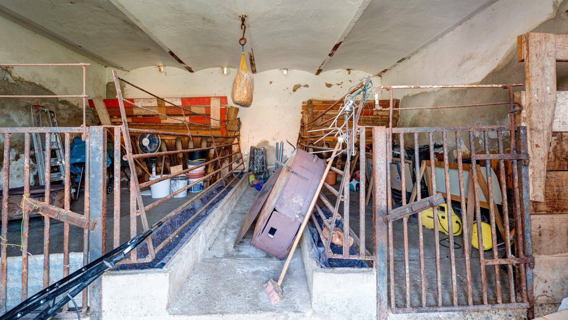Alter Stallbereich mit Metallgittern und verschiedenen gelagerten Gegenständen.