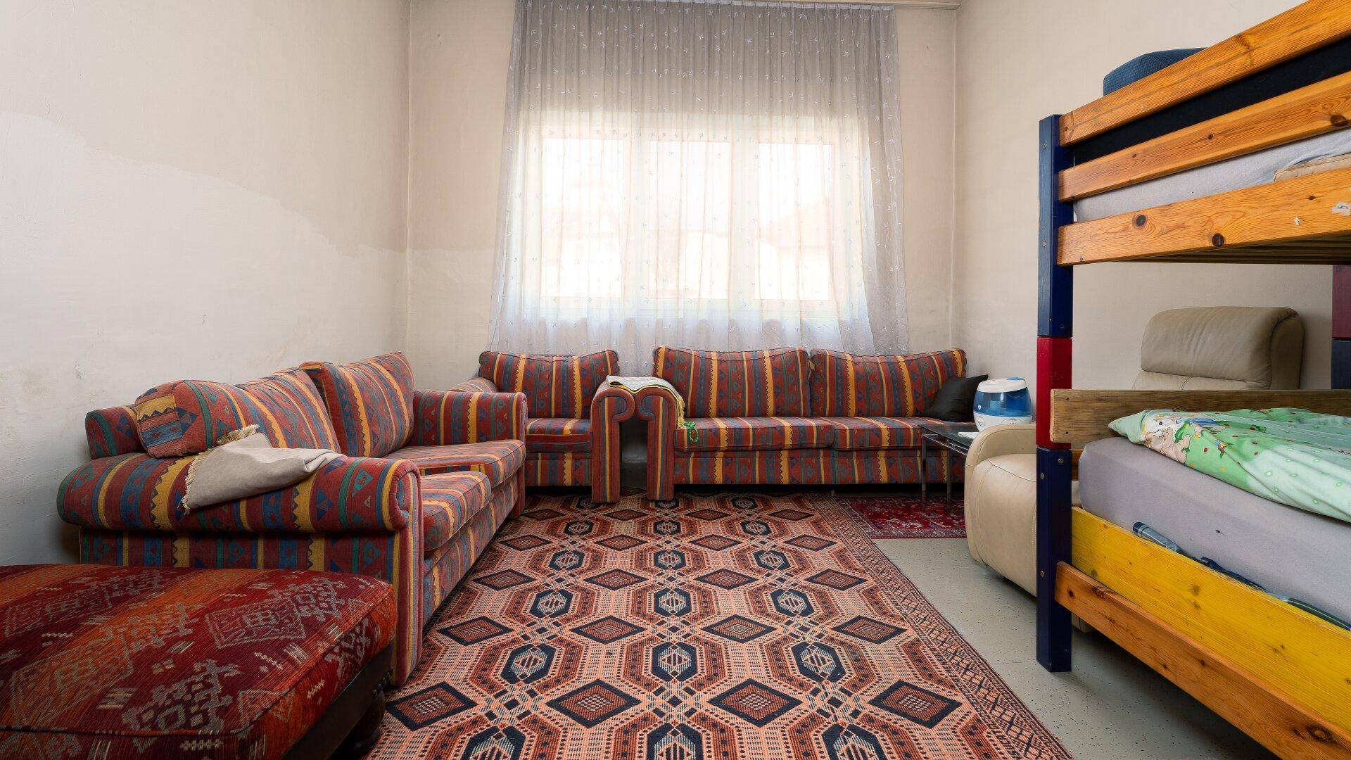 Einfaches Wohnzimmer mit bunten Sofas, Etagenbett und gemustertem Teppichboden.