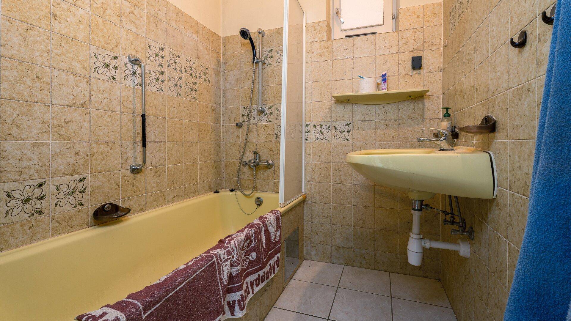 Badezimmer mit gelber Badewanne, Duschvorrichtung und gefliesten Wänden im 80er-Jahre-Stil.