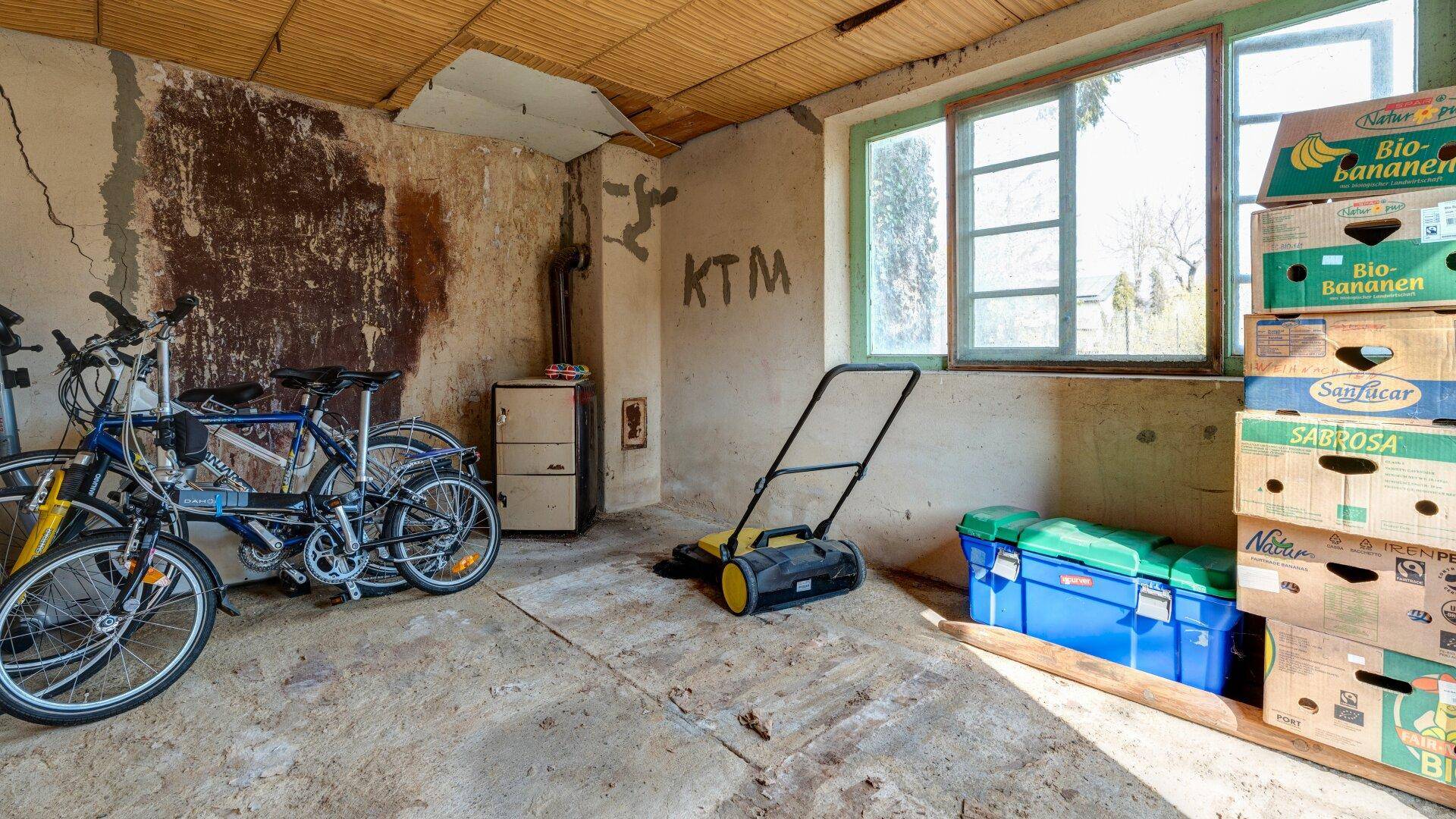 Lagerraum mit Fahrrädern, Rasenmäher und Kartons, beleuchtet durch ein Fenster.
