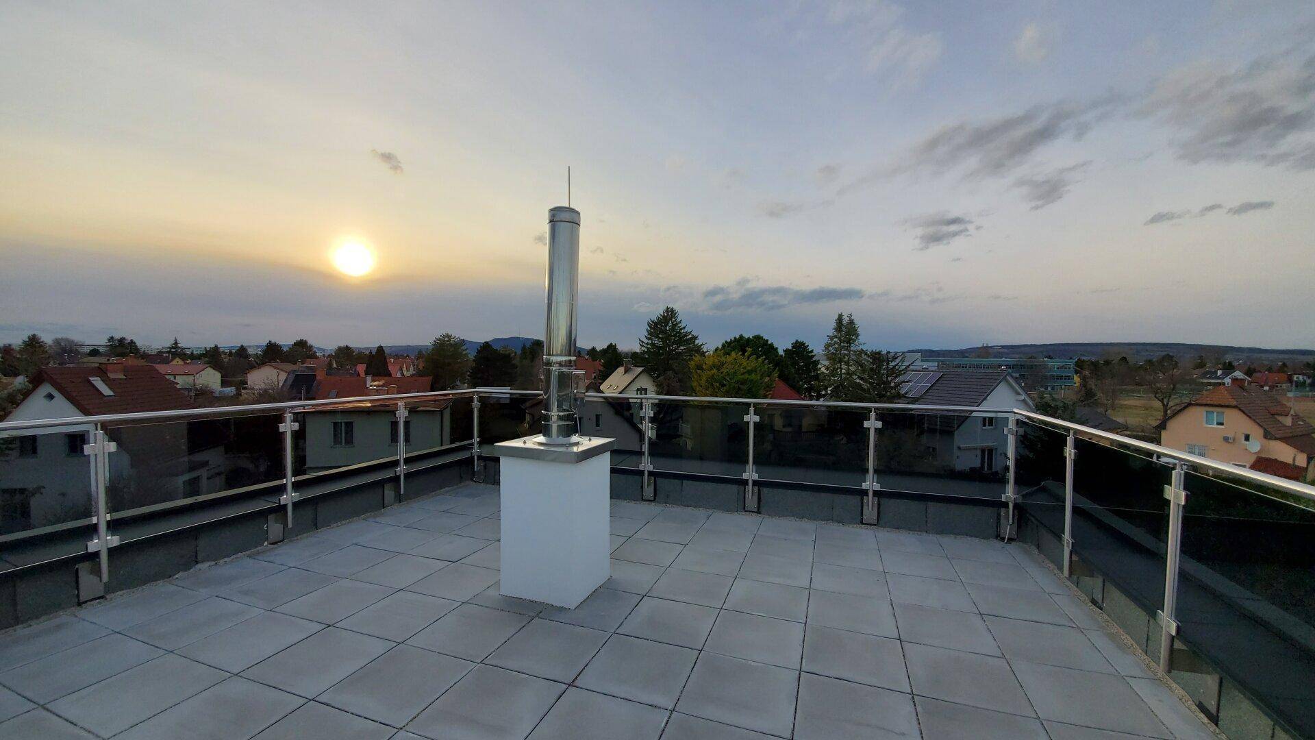 Weitläufige Dachterrasse mit Fliesenboden und Glasgeländer, weiter Blick bei Sonnenuntergang.