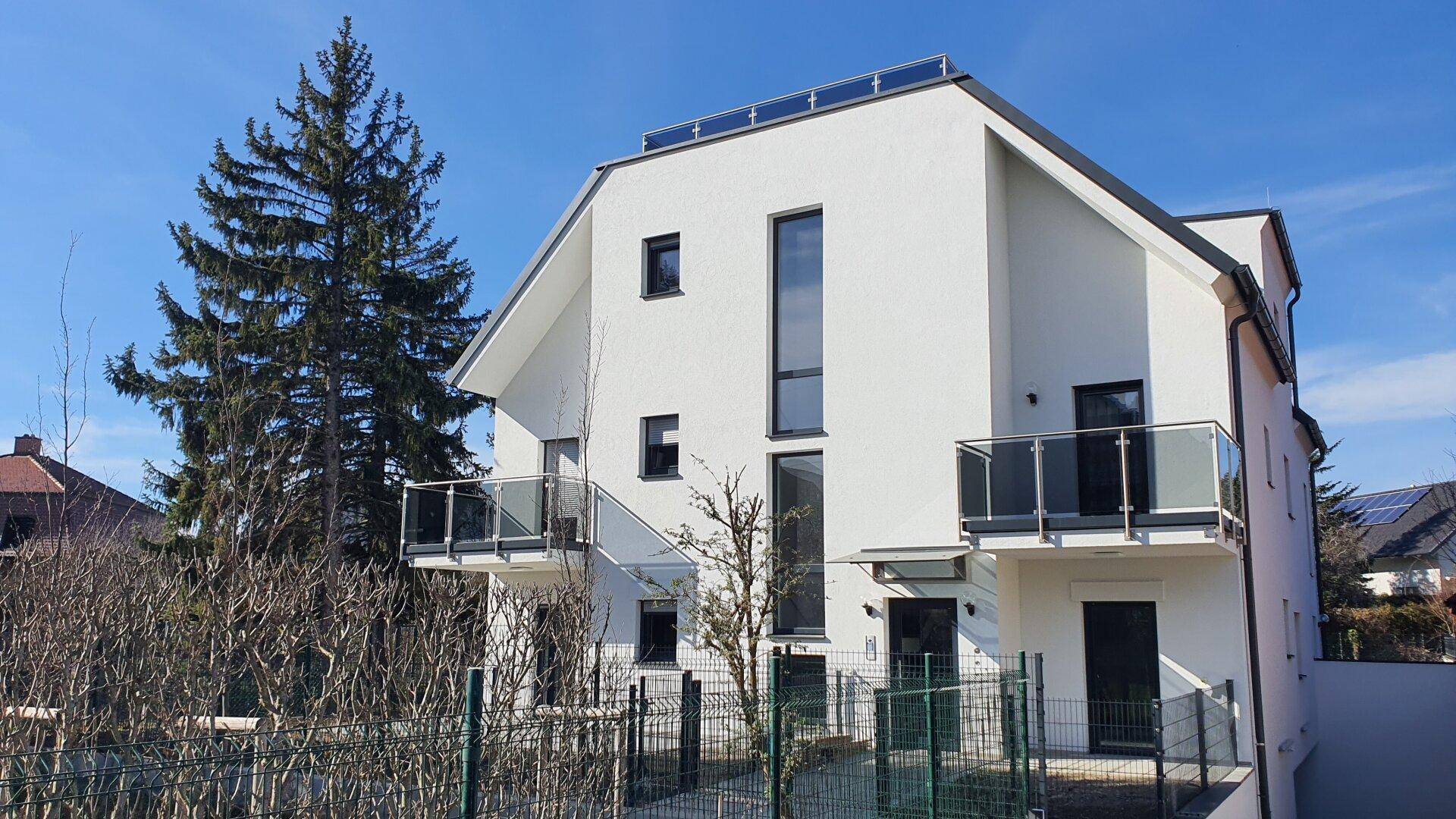 Moderne weiße Fassade eines Neubaus mit Balkonen und großen Fenstern unter blauem Himmel.