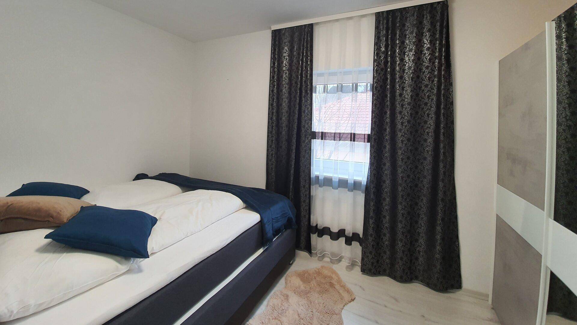 Schlafzimmer mit Doppelbett, Fenster und dunklen Vorhängen, heller Laminatboden.