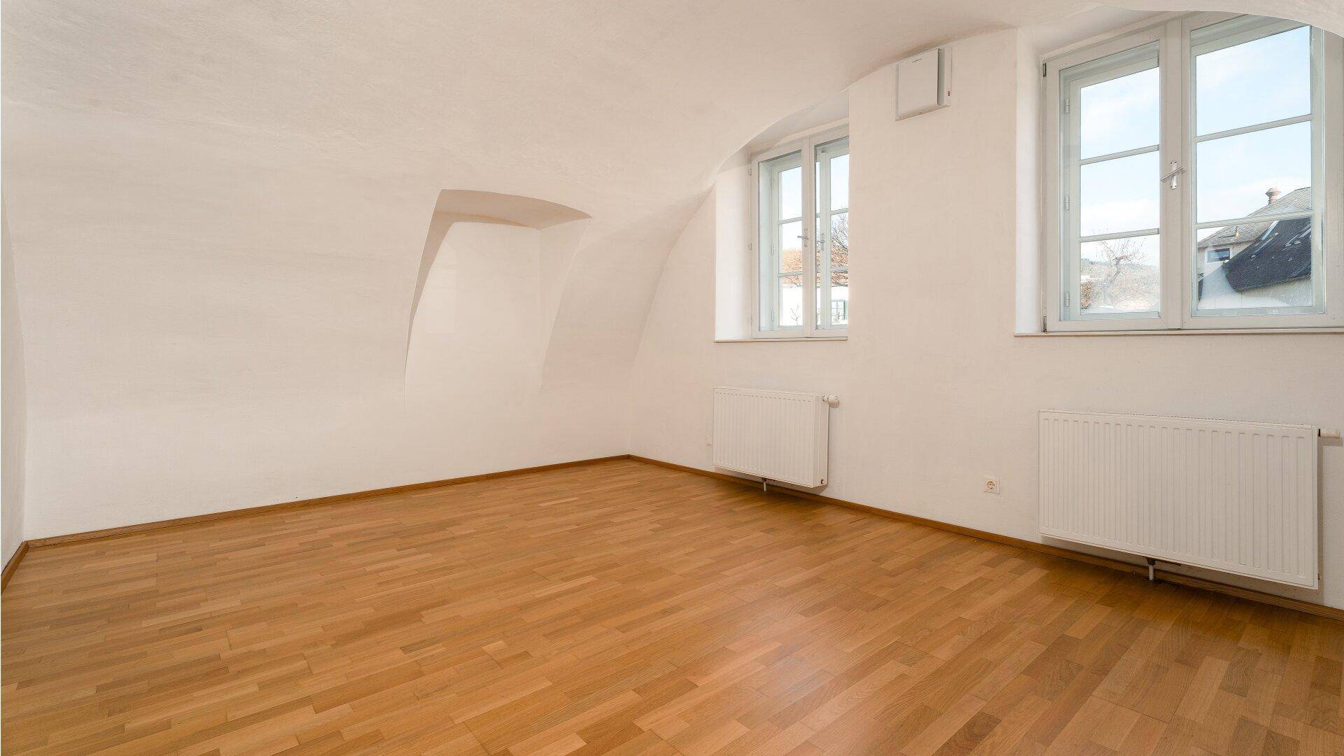 Leeres Zimmer mit Parkettboden, zwei Fenstern und gewölbter Decke, ideal als Schlaf- oder Wohnzimmer.