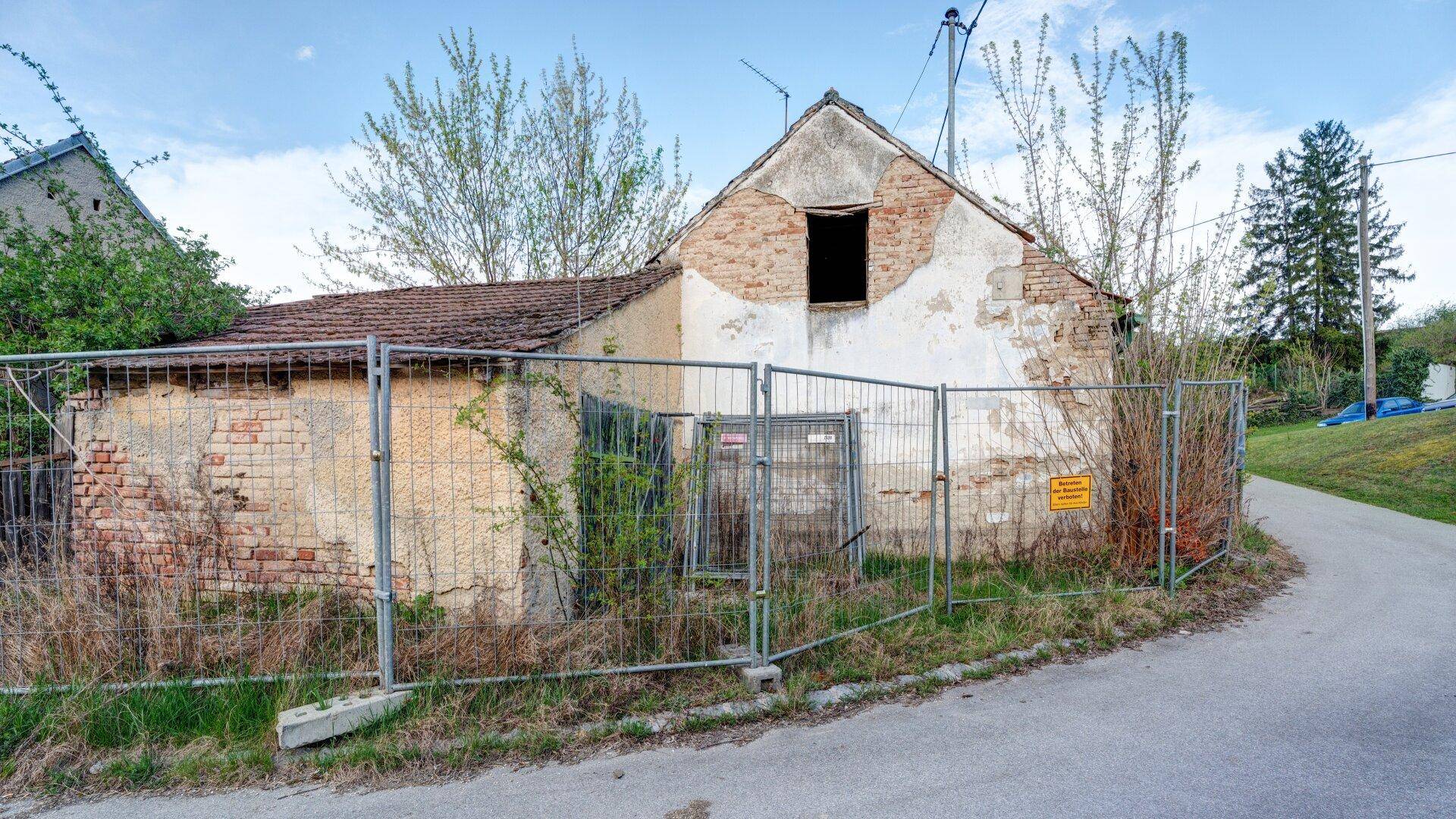 Altes, sanierungsbedürftiges Haus mit bröckelndem Putz und Bauzaun an einer ländlichen Straße.