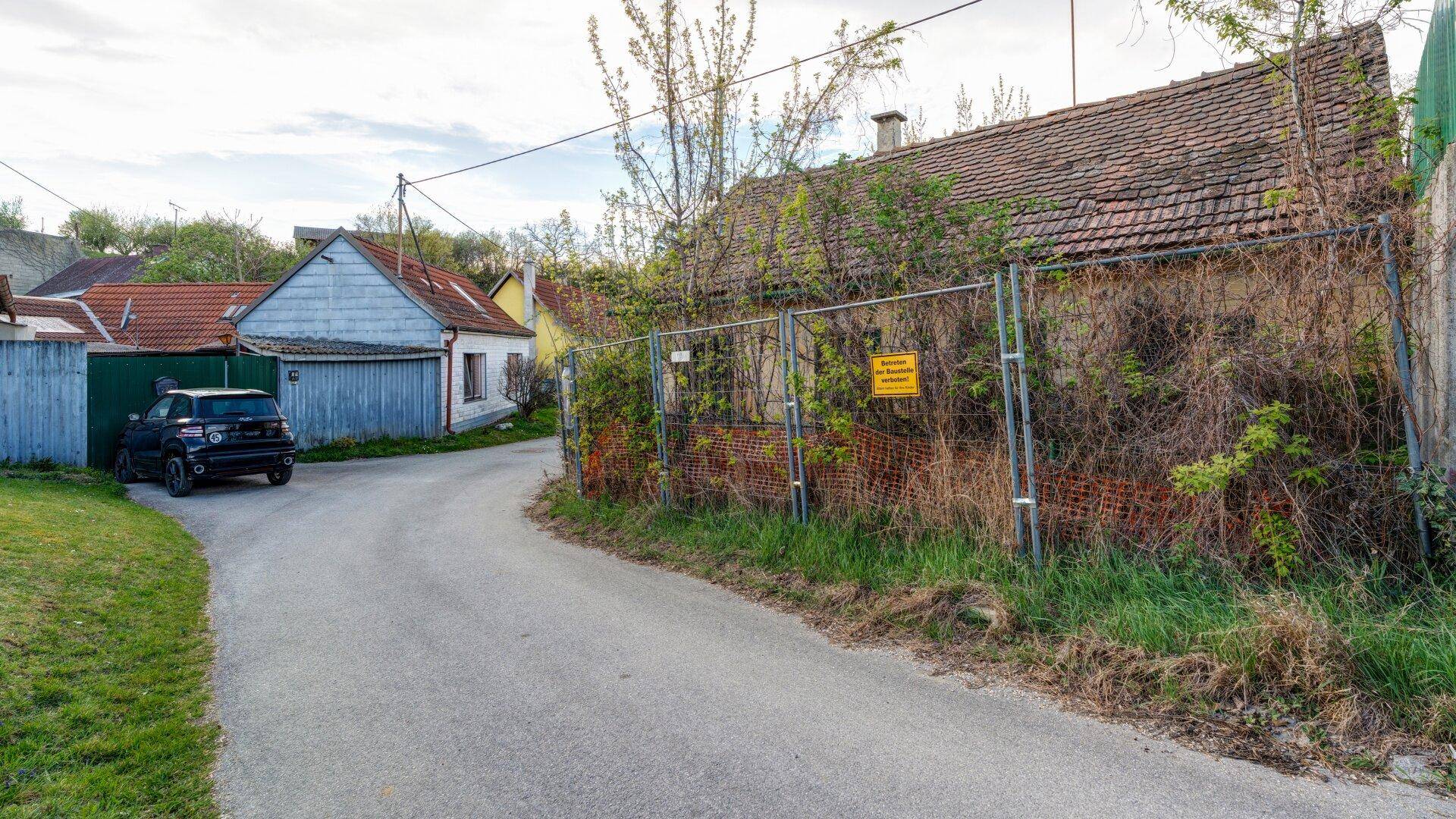 Verwildertes Grundstück mit altem Haus und Bauzaun entlang einer ruhigen Nebenstraße in ländlicher Gegend.