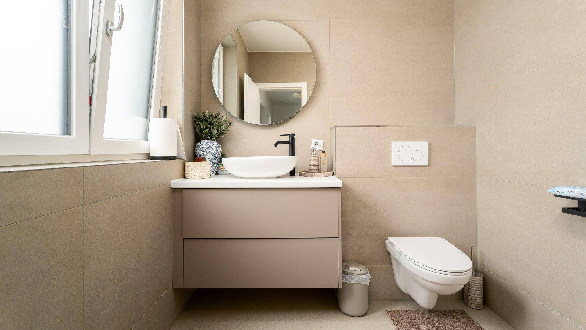 Modernes Gäste-WC mit beigen Fliesen, wandhängendem WC und rundem Spiegel.