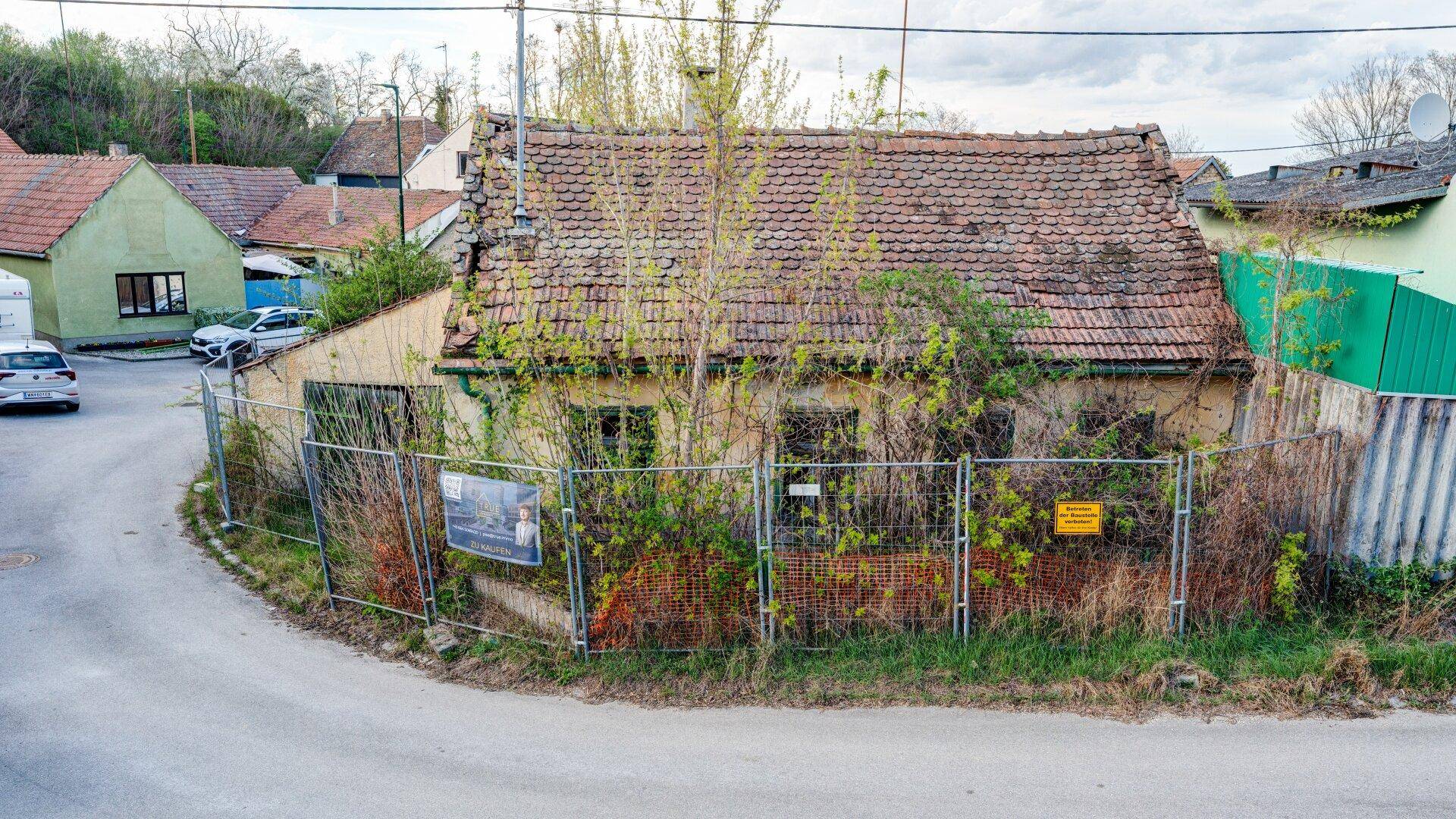 Blick auf ein stark renovierungsbedürftiges Gebäude mit überwuchertem Garten und Bauzaun an der Straße.