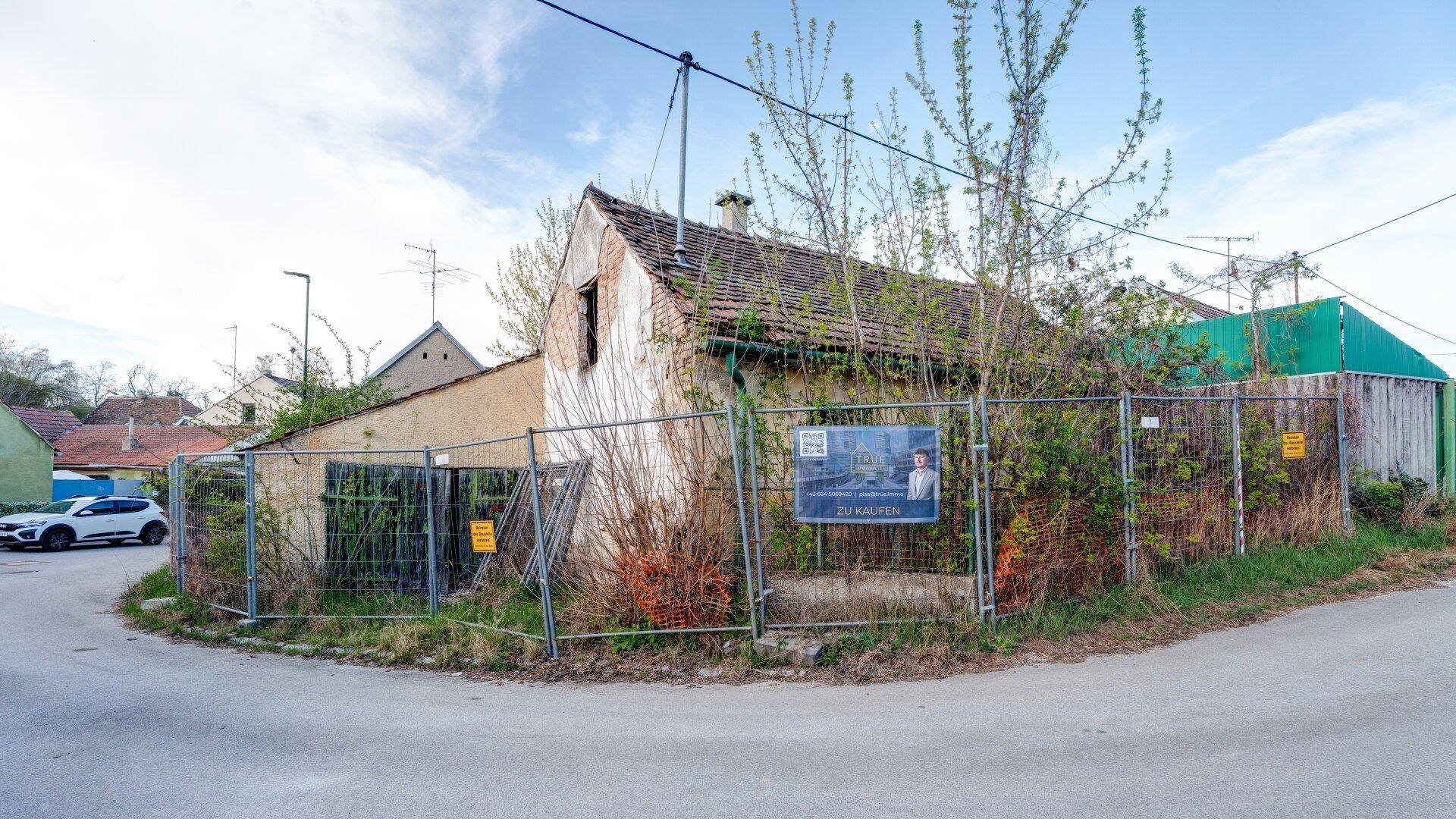 Eckgrundstück mit altem Haus, das zum Verkauf steht, umgeben von einem Bauzaun und Bäumen.