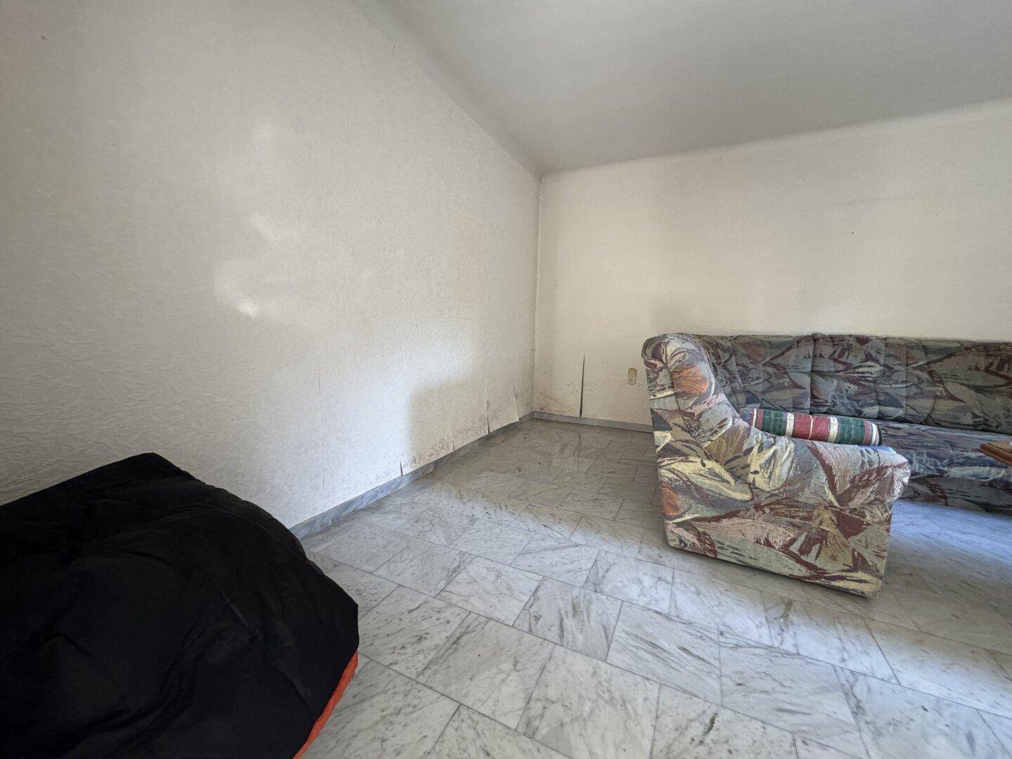 Einfacher Wohnbereich mit Fliesenboden und einem Sofa, das auf Renovierung wartet.