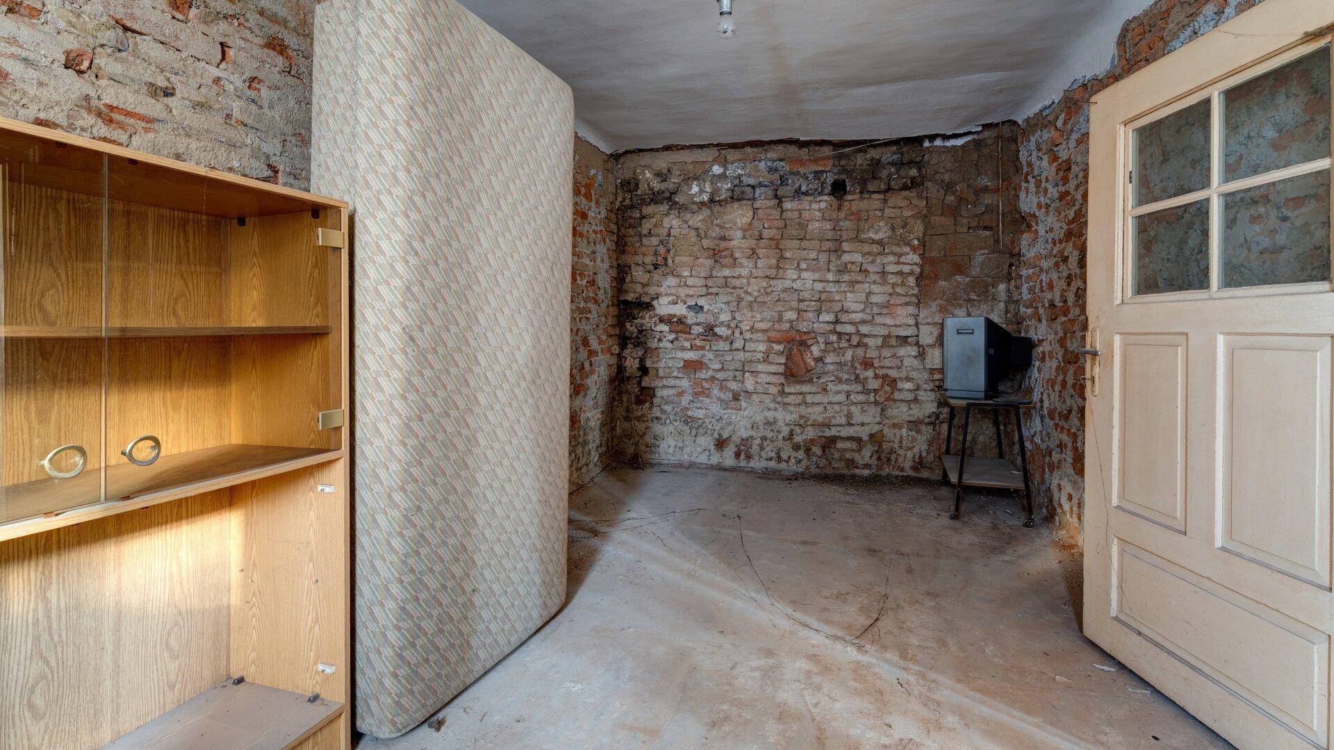 Schlafzimmer im Rohbau mit freiliegenden Ziegelwänden, Matratze und Blick auf eine weitere Tür.