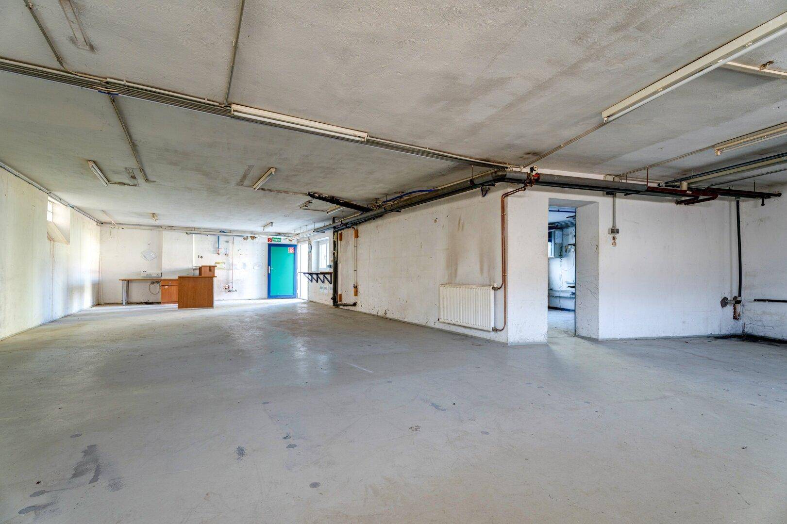 Großer, leerer Kellerraum mit Betonboden und sichtbaren Leitungen an der Decke.