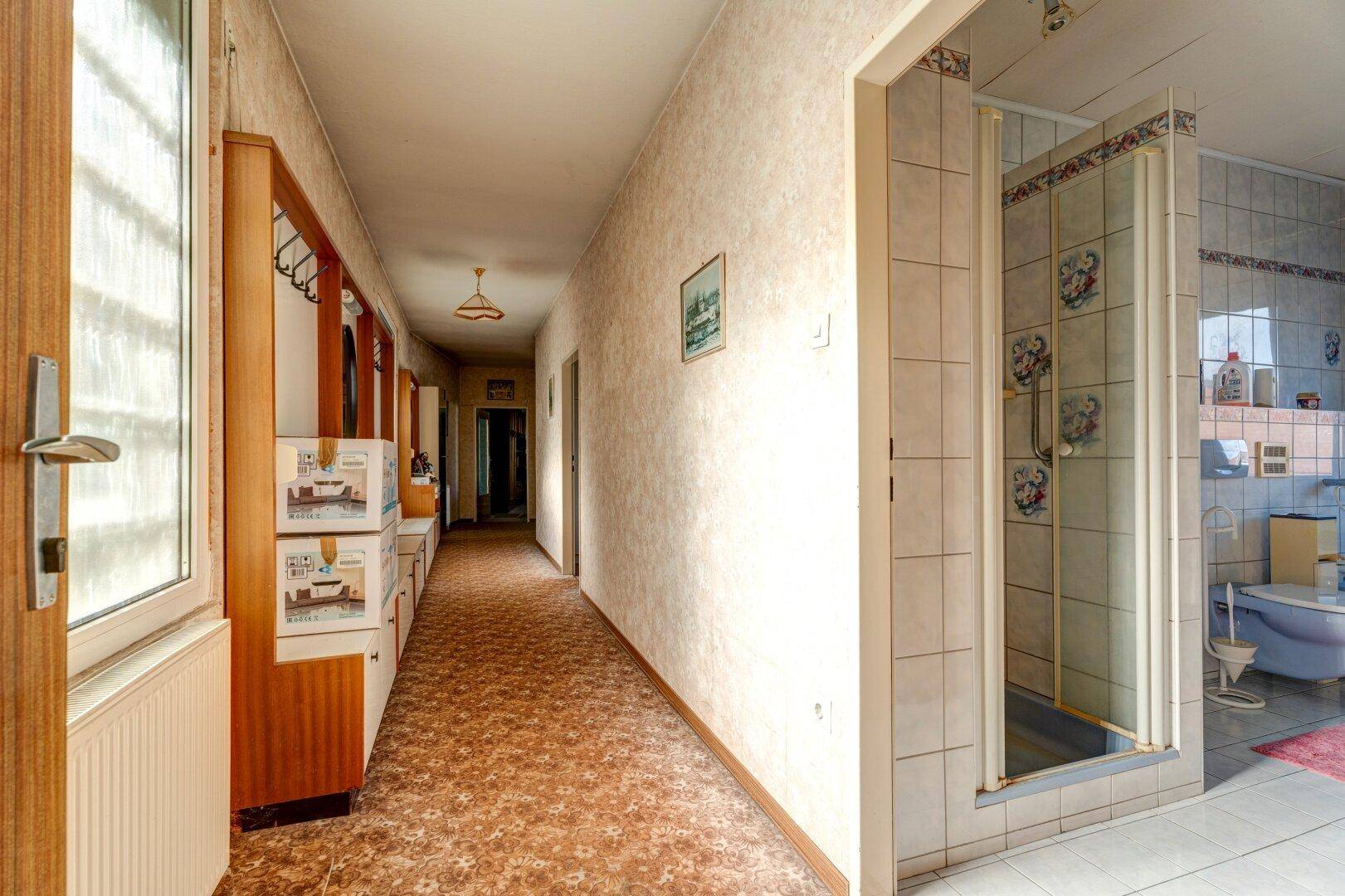 Langer Flur mit gemusterter Auslegware und Blick in ein gefliestes Badezimmer mit Duschkabine.
