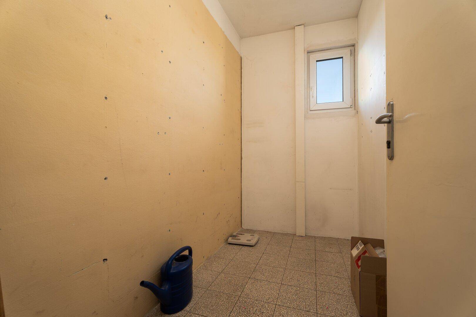 Abstellraum mit kleinem Fenster, Fliesenboden und gelben Wänden.