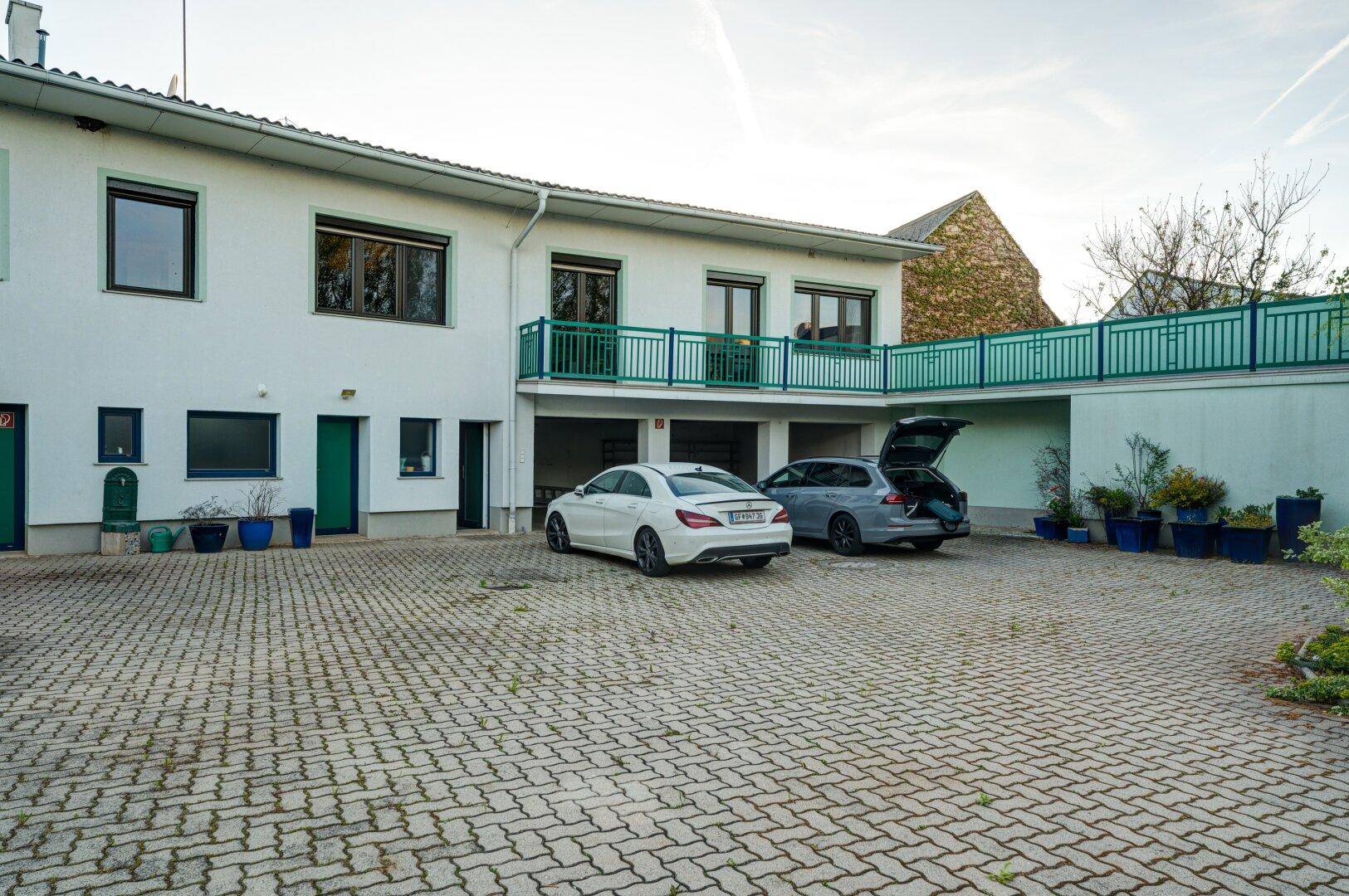 Außenansicht des Gebäudes mit gepflastertem Parkplatz und Autos sowie einem grünen Balkon.