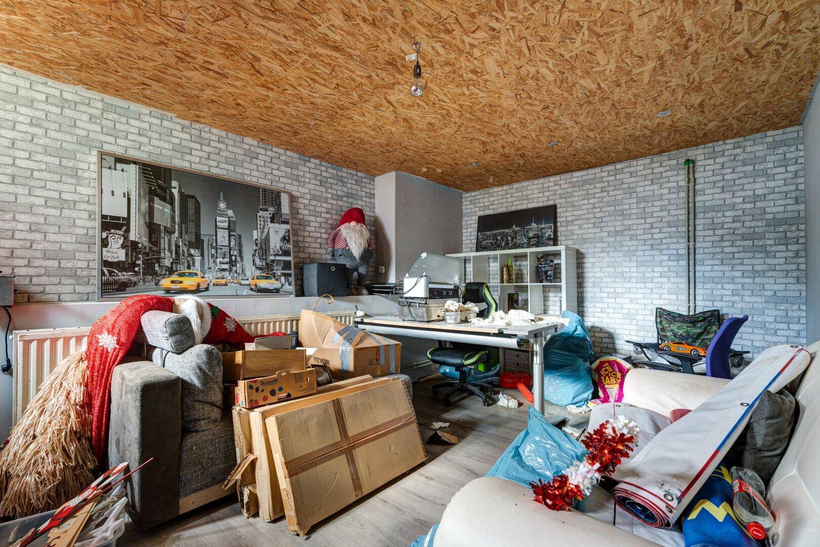 Ein unaufgeräumter Raum mit heller Ziegelmuster-Tapete, einer Holzspanplatten-Decke und verschiedenen Möbeln und Kartons.