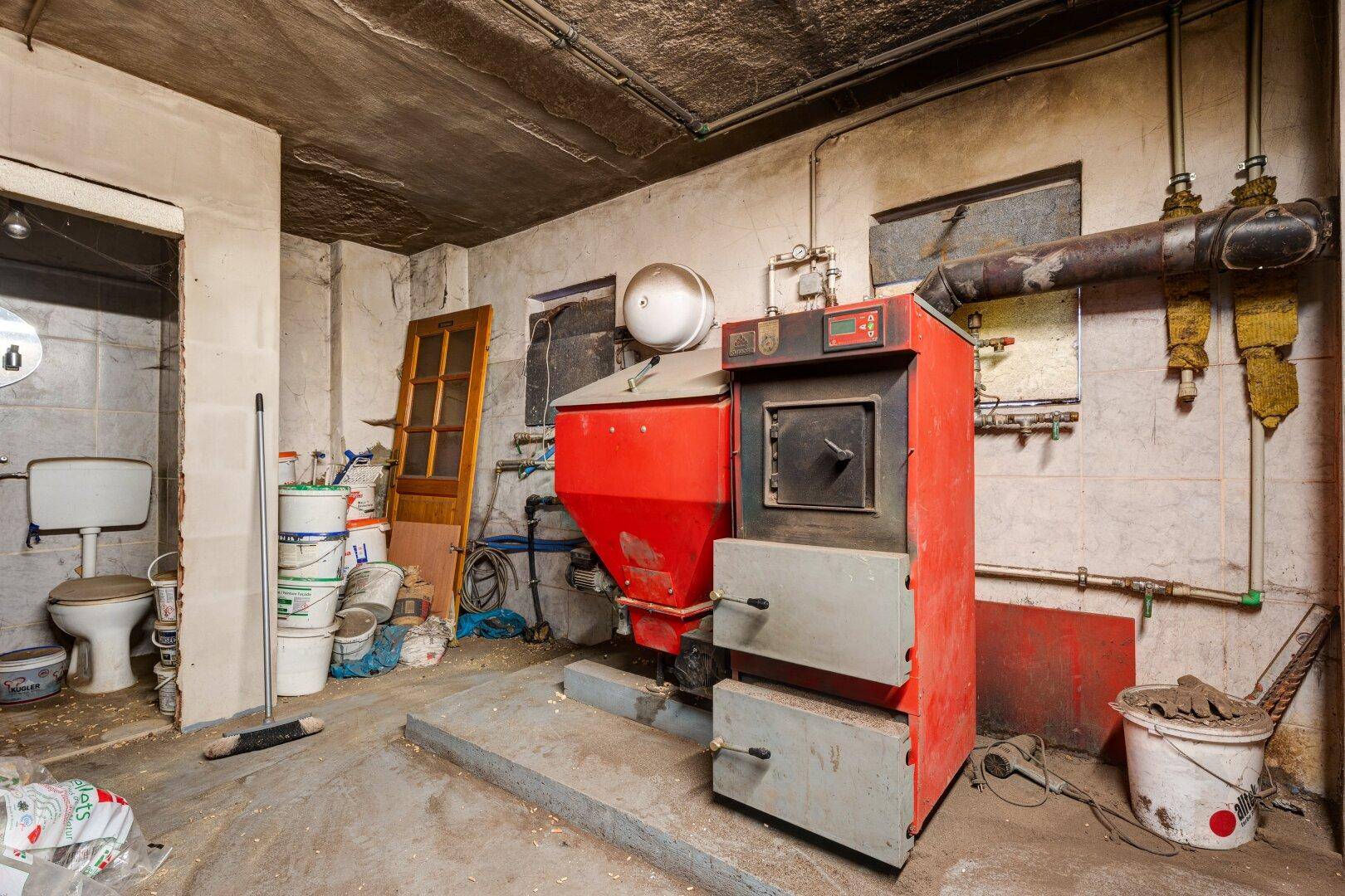 Ein Hauswirtschaftsraum oder Keller mit einem großen roten Heizkessel, Rohren und einer Toilette im Hintergrund.