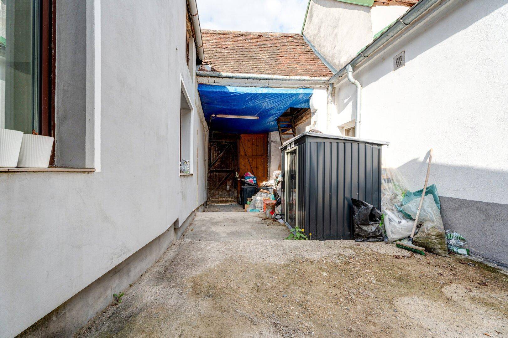 Eine weitere Ansicht des betonierten Außenwegs zwischen Gebäuden, mit einem kleinen Schuppen und einer Holztür am Ende.