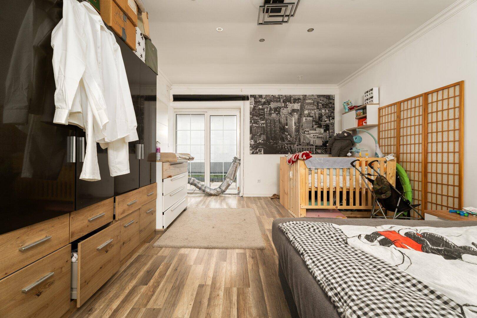 Ein Schlafzimmer mit einem Doppelbett, einem Babybett, einem großen Kleiderschrank und einer Schiebetür nach draußen.