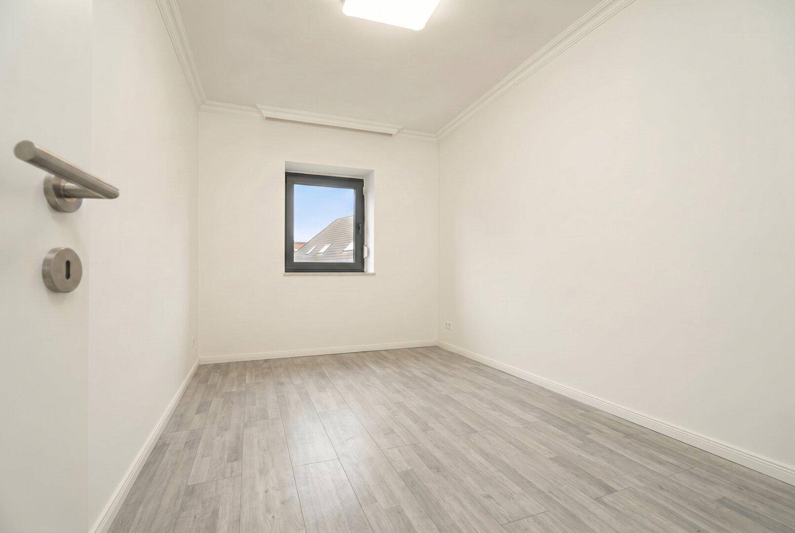 Ein leerer Raum mit weißen Wänden, hellgrauem Laminatboden und einem einzelnen Fenster.