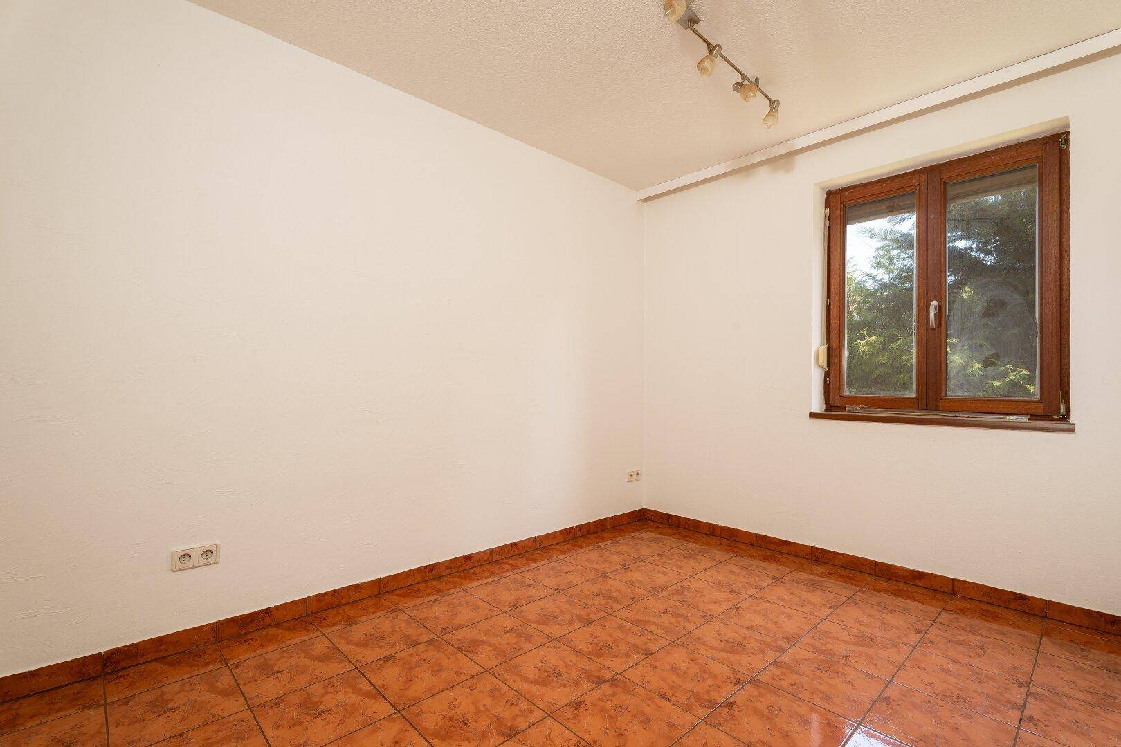 Leeres Zimmer mit Terrakotta-Fliesenboden und einem Fenster mit Blick ins Grüne.
