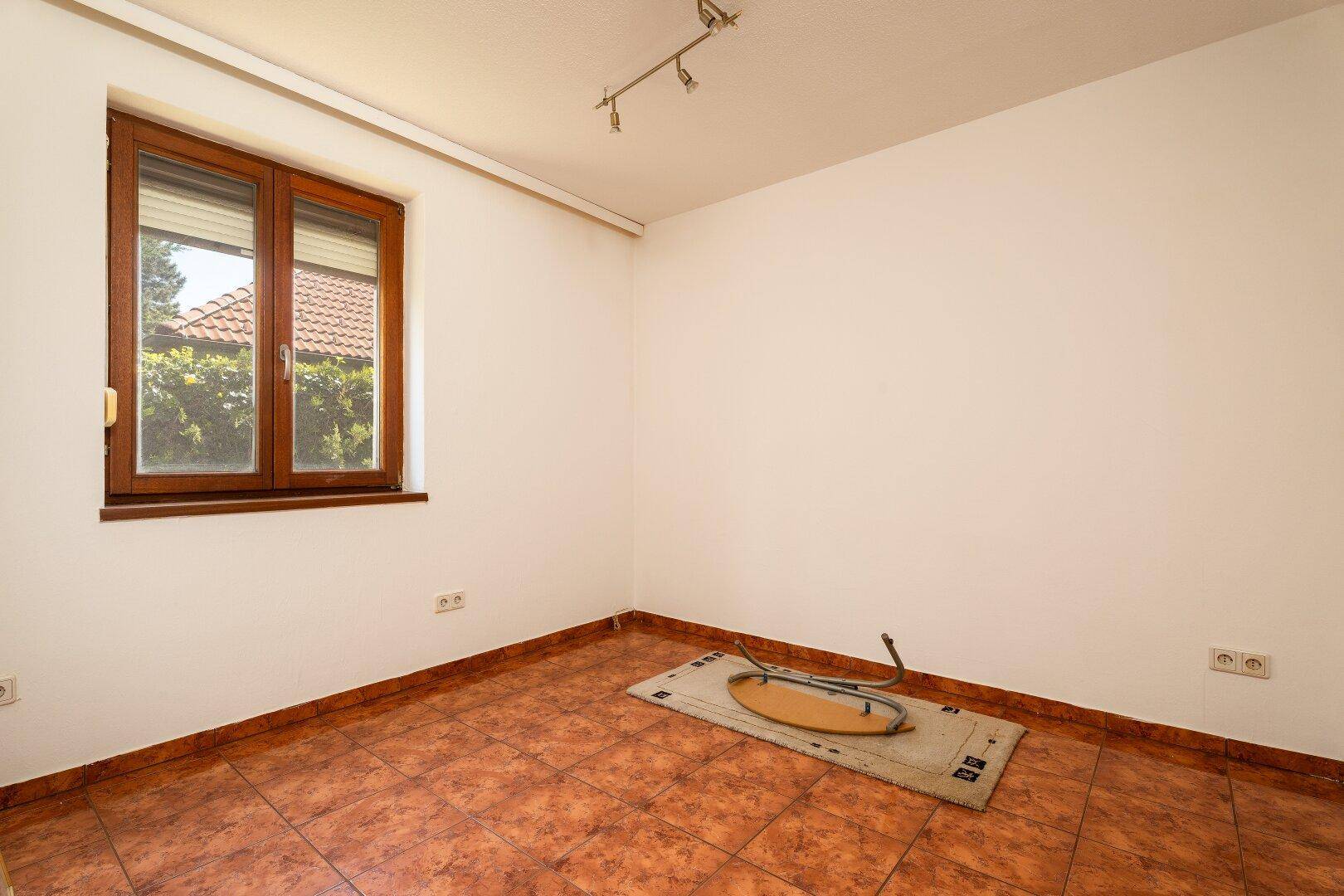Helles, leeres Zimmer mit Terrakotta-Fliesenboden und Fenster mit Rollladen.