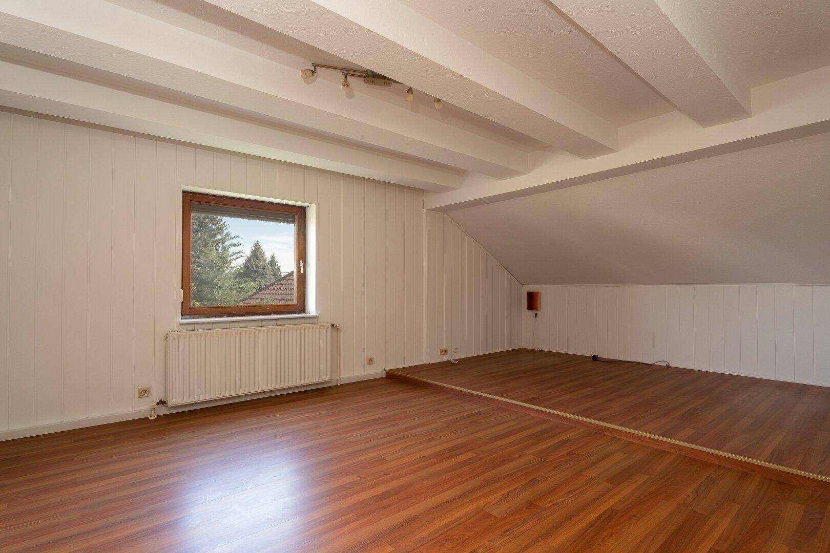Geräumiges Dachzimmer mit Holzboden, sichtbaren Deckenbalken und Fenster.