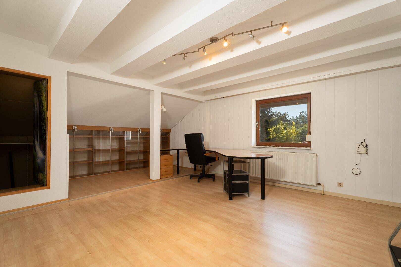Helles Dachgeschossbüro mit Holzboden, sichtbaren Deckenbalken und großem Schreibtisch.