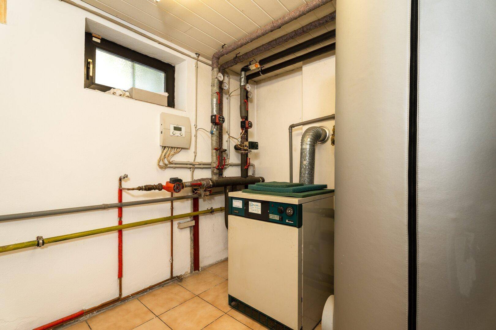 Heizungsraum im Untergeschoss mit Heizkessel, Rohren und kleinem Fenster.