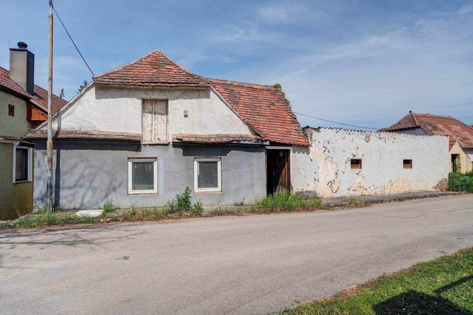 Seitenansicht des alten Hauses mit verputzter grauer Fassade, rotem Ziegeldach und einer baufälligen Mauer.
