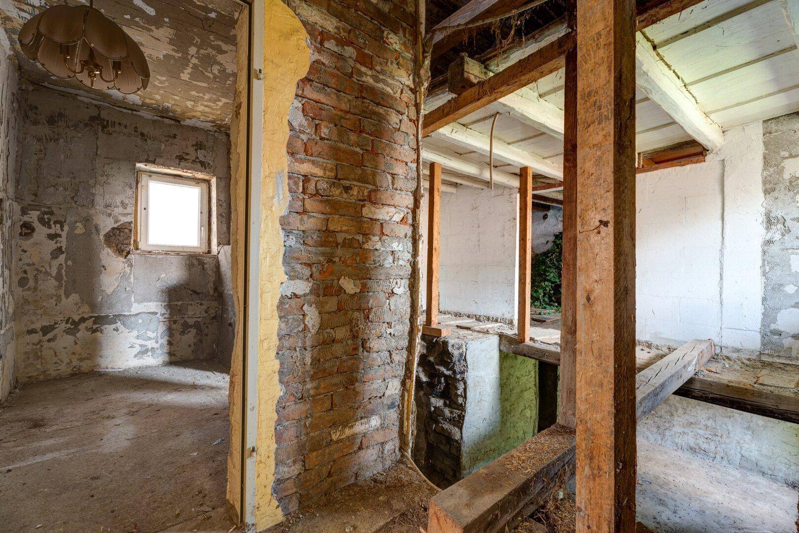Innenansicht mit freiliegenden Ziegelwänden, Holzbalken und einem Fenster in einem stark renovierungsbedürftigen Zustand.